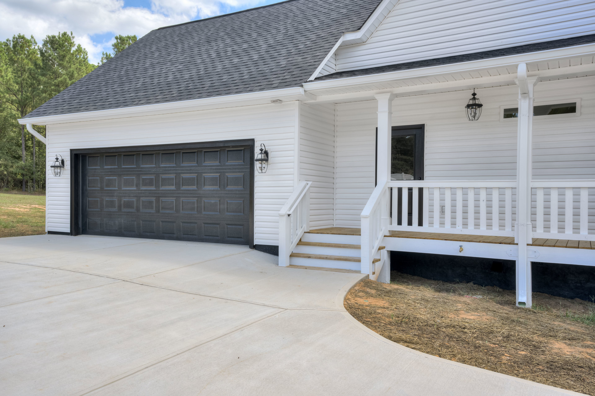 Two-story home with white siding, black garage door with white trim, concrete driveway, front porch with white railing, black front door, and straw-covered ground.
