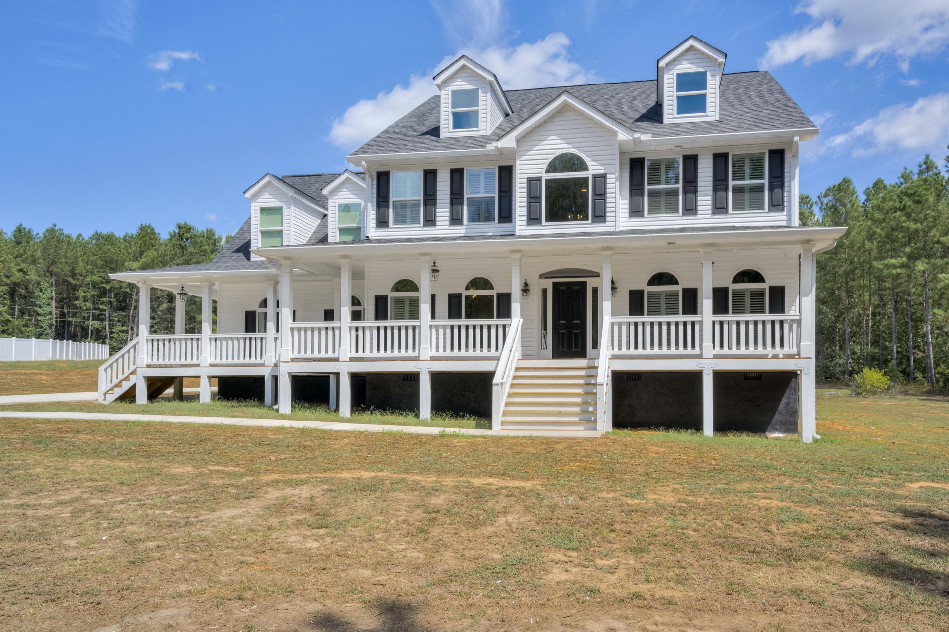 White two-story house with wide front porch, black door, wooden railing, large windows, and expansive green lawn with stone steps.
