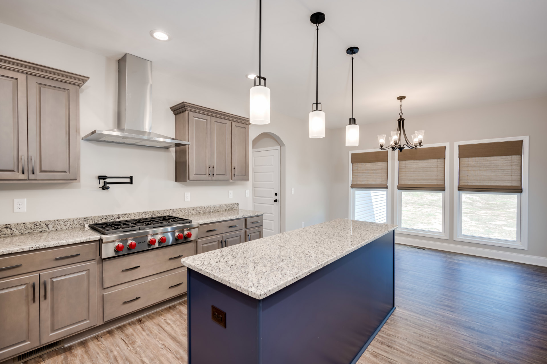 Blue kitchen island with white countertop, wood flooring, white cabinetry, stainless steel stove with red knobs, window with brown shade, and black support pole