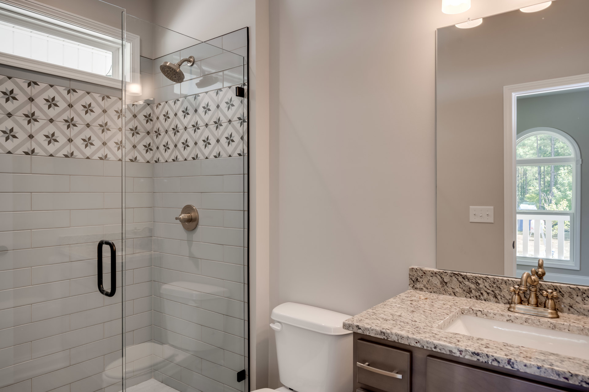 Bathroom with glass shower enclosure, marble-topped white toilet, modern sink and faucet on stone counter, window overlooking trees, tile walls, chrome plumbing fixtures