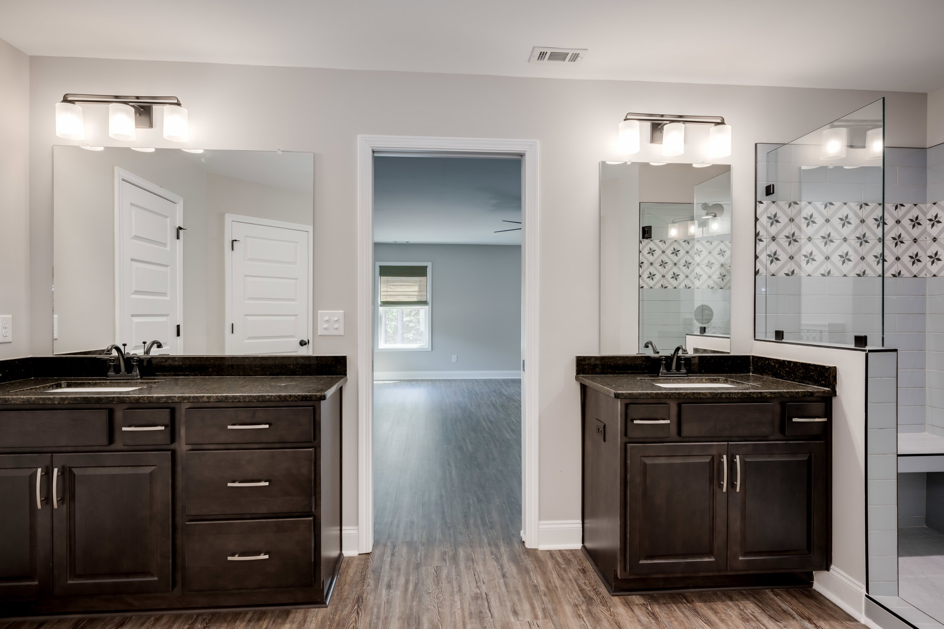 Bathroom with double sinks set in a black countertop, two rectangular mirrors above, white cabinetry with silver handles, wood flooring, white door with black handle, window