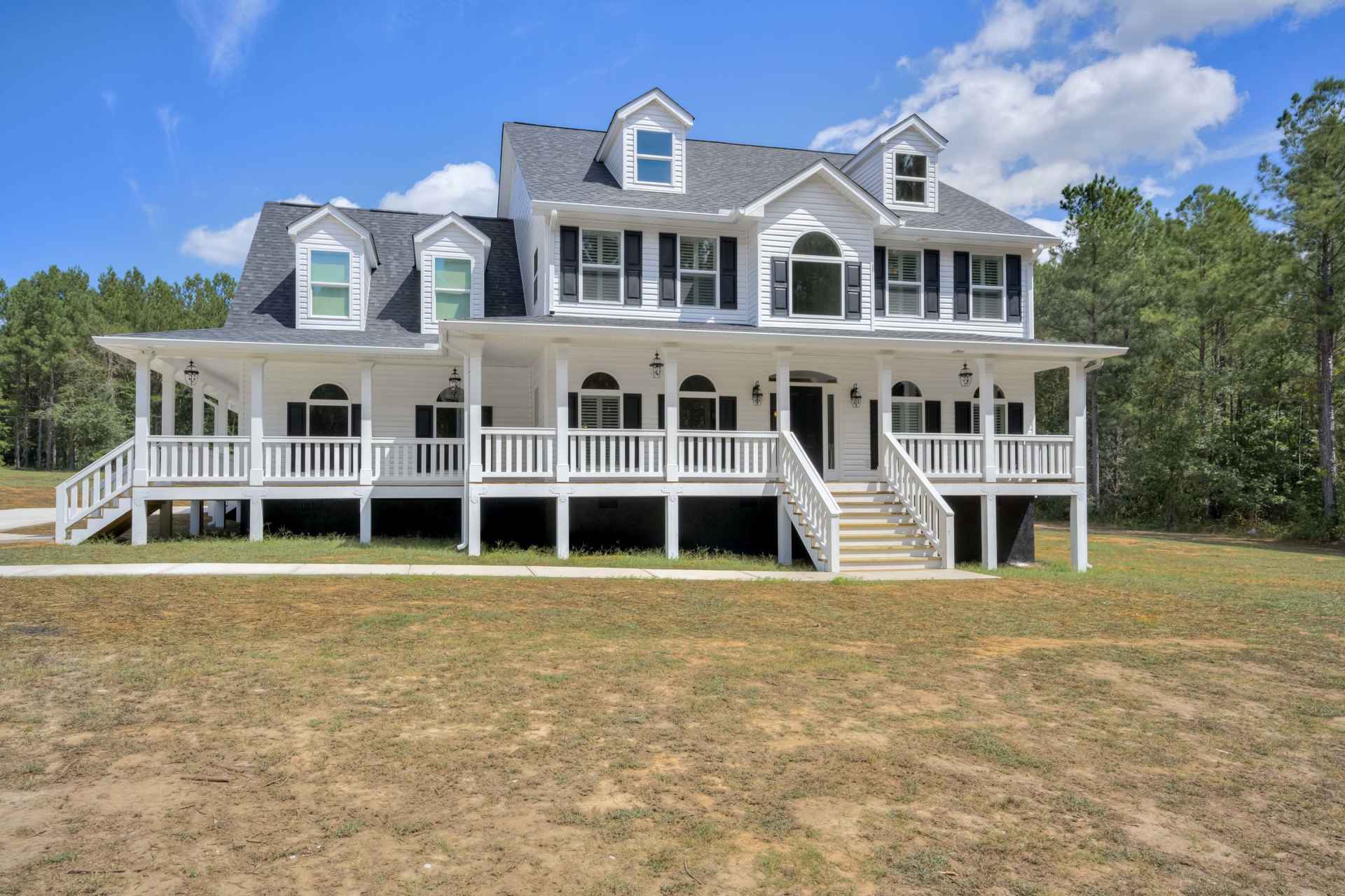 White two-story house with wide covered front porch, white railings and columns, expansive green lawn, multiple windows with white trim, and exterior staircase leading to porch.