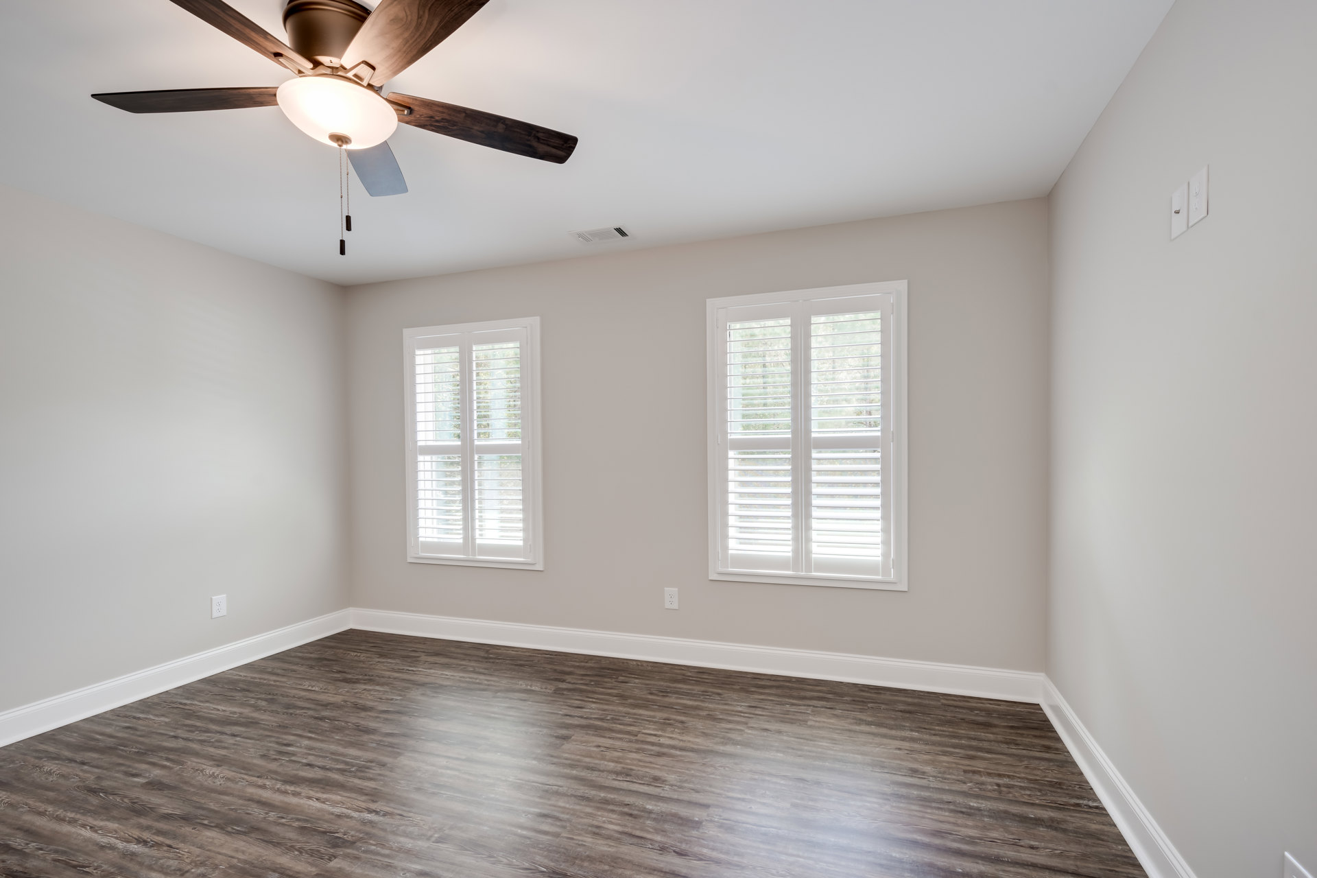 Ceiling fan with light fixture mounted on white plaster ceiling, wood flooring with white baseboards, windows fitted with blinds, and a white electrical outlet with silver plug
