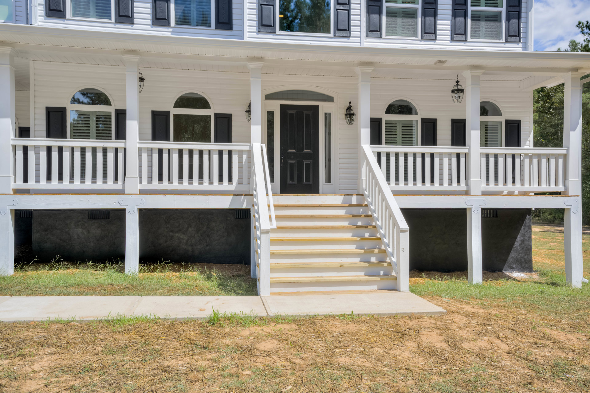 White house exterior with black shutters, black front door, white porch with baluster railings, and white stairs leading up from grass and dirt yard