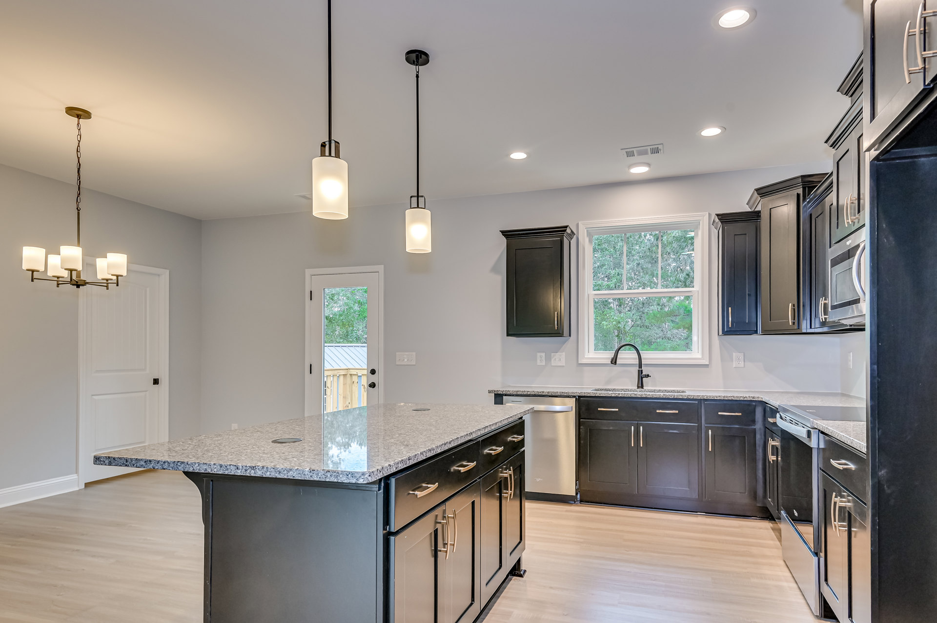 Spacious kitchen featuring a marble-topped island, white cabinetry, stainless steel appliances, black door with handle, window above sink overlooking trees, and modern pendant