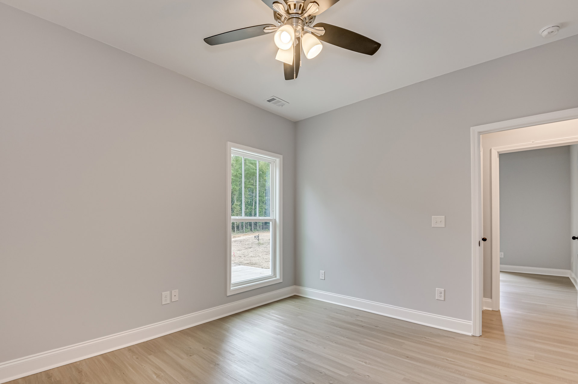 Ceiling fan with light fixture mounted above wood laminate flooring, large window with view of trees and outdoor sign, white plaster walls with crown molding