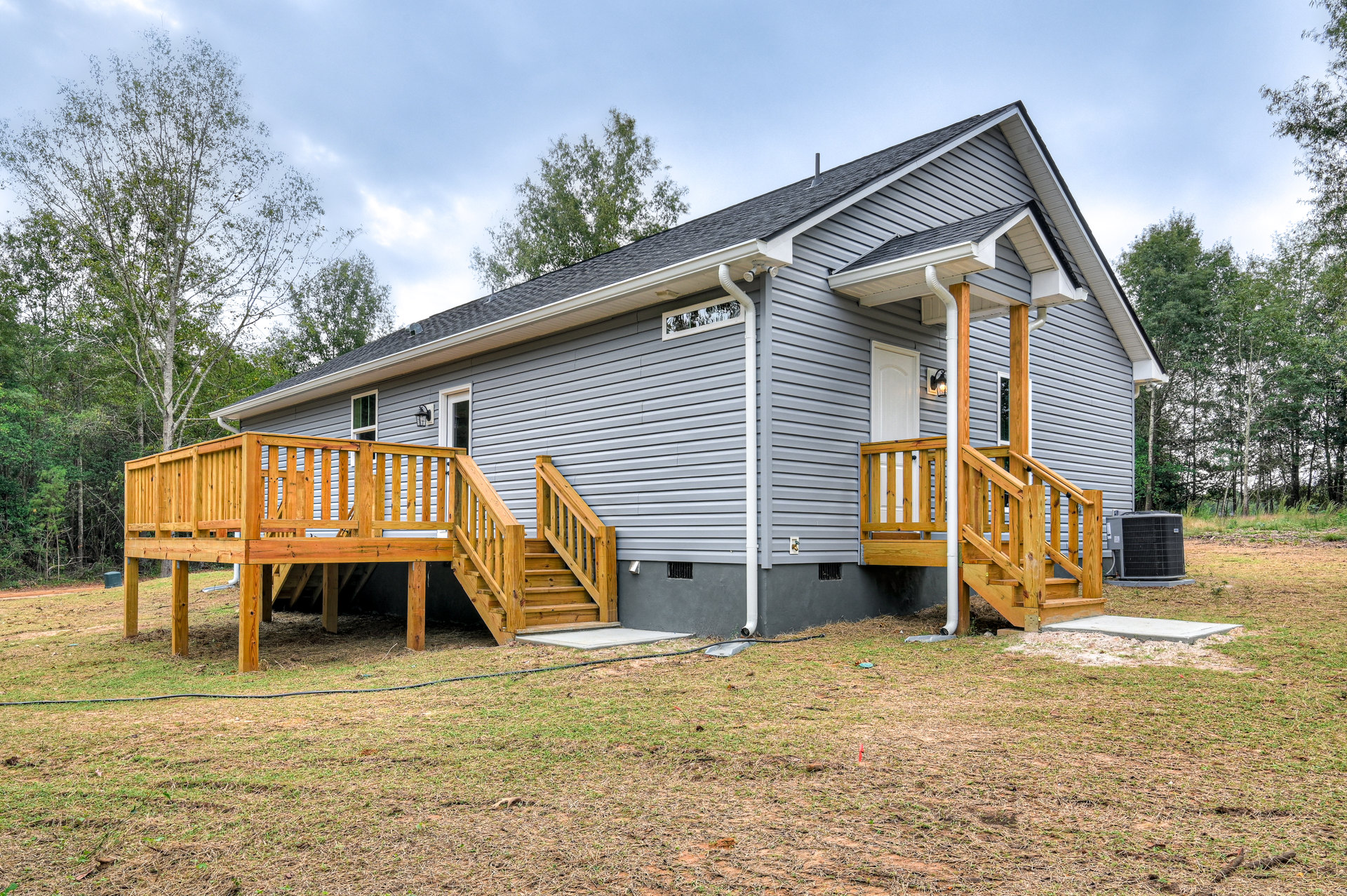Wooden porch with railing and stairs leading to entry, grey metal gutter attached above, surrounded by grass, trees, and cloudy sky