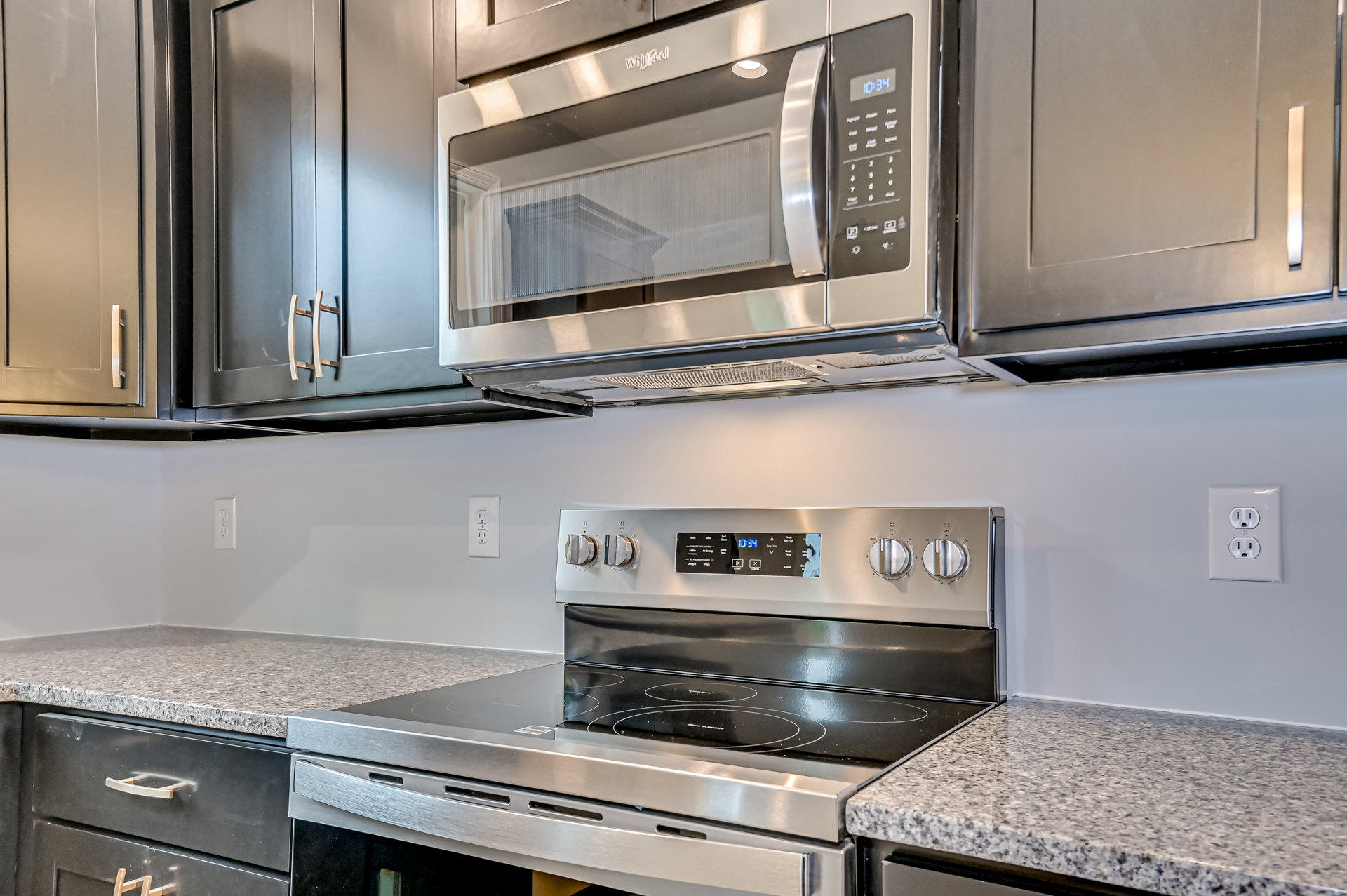 White kitchen with stainless steel microwave, stove, and digital clock on black panel, surrounded by light cabinetry and stone countertops