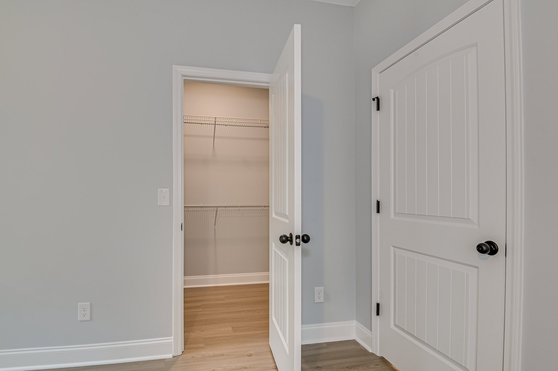 White closet door with matte black knob open to reveal metal shelving against white walls, wood flooring, and nearby light switch.