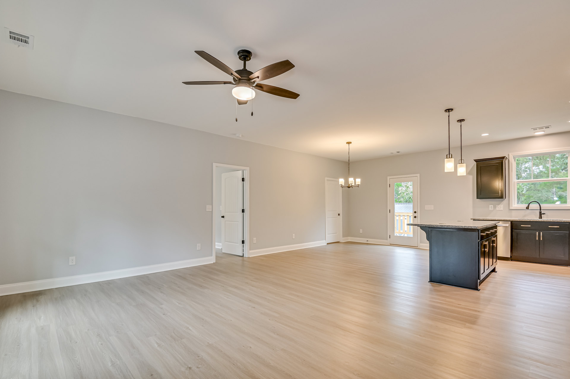 Open concept kitchen and dining area with a black marble-topped island, brown cabinets, laminate wood flooring, ceiling fan with light, white door featuring black knobs and window.