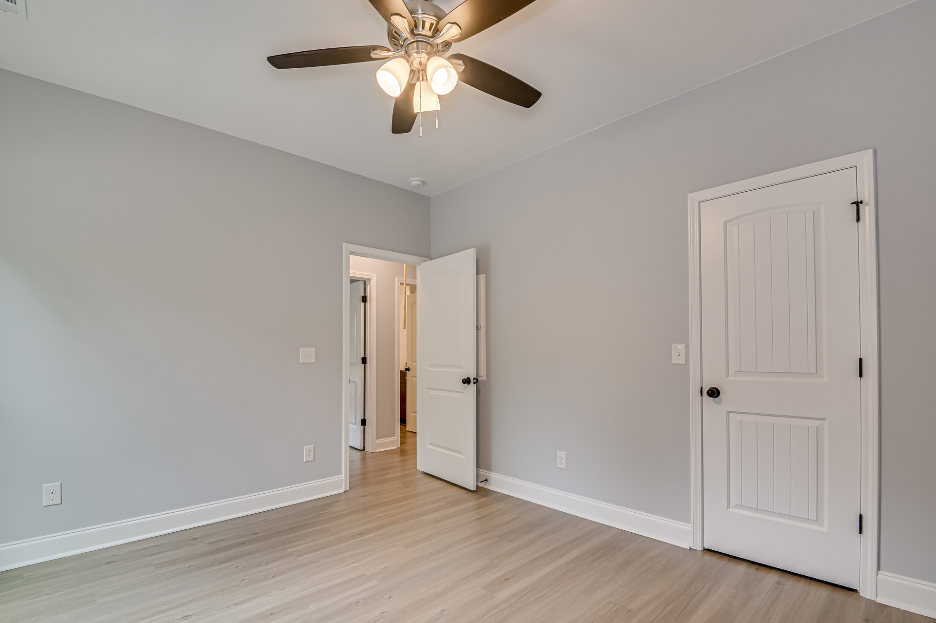 Ceiling fan with integrated lights mounted on white ceiling, white paneled door with black knob opening to neutral painted wall, light wood flooring and simple base molding