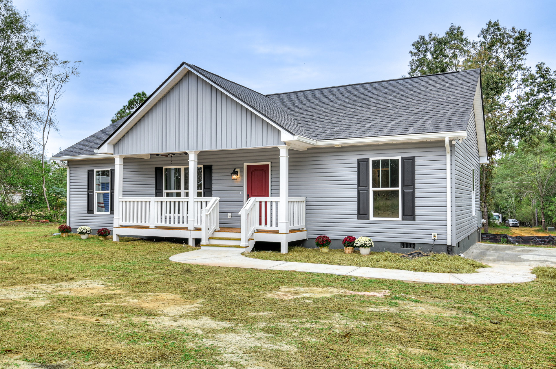 Two-story cottage with gray siding, red front door, white porch railing, broken window glass, manicured lawn, potted flowers, and mature trees in the yard
