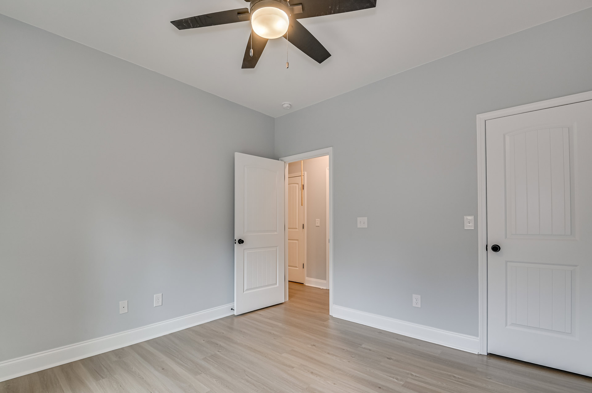 Ceiling fan with integrated light fixture above wood flooring, white paneled doors with contrasting round and black door knobs, neutral walls, and simple trim in a residential room