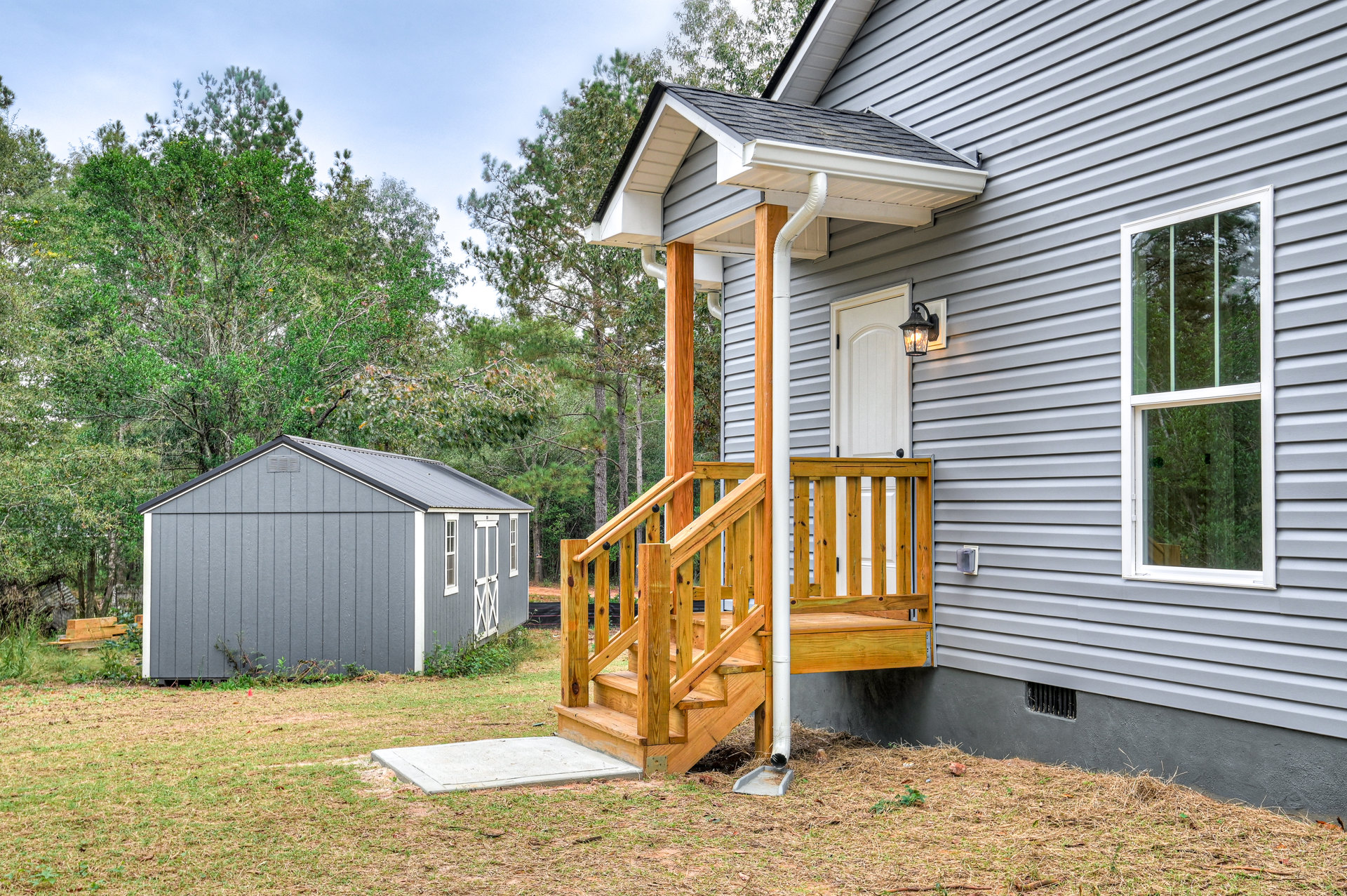 Gray siding house with white trim, wooden porch featuring white columns and double doors, concrete slab set in grassy yard, gray shed with window, mature trees visible through