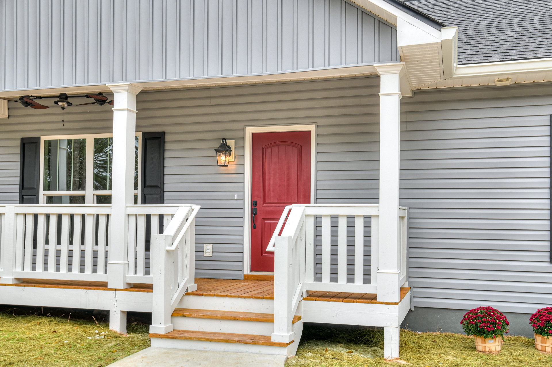 Front porch with white railing, red door, hanging basket of red flowers, wall-mounted light fixture, gray siding, steps leading to entry, green grass in yard