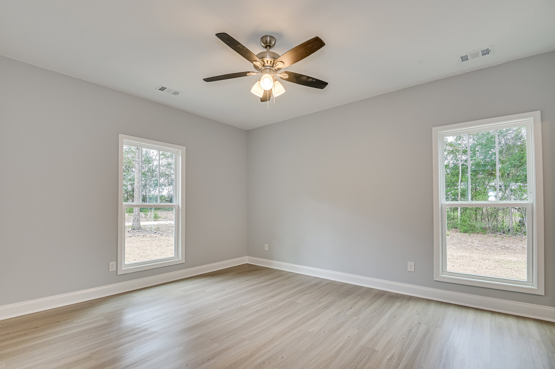 Ceiling fan with light fixture centered in a room featuring wood flooring, white baseboards, large windows overlooking trees, and a wall vent.
