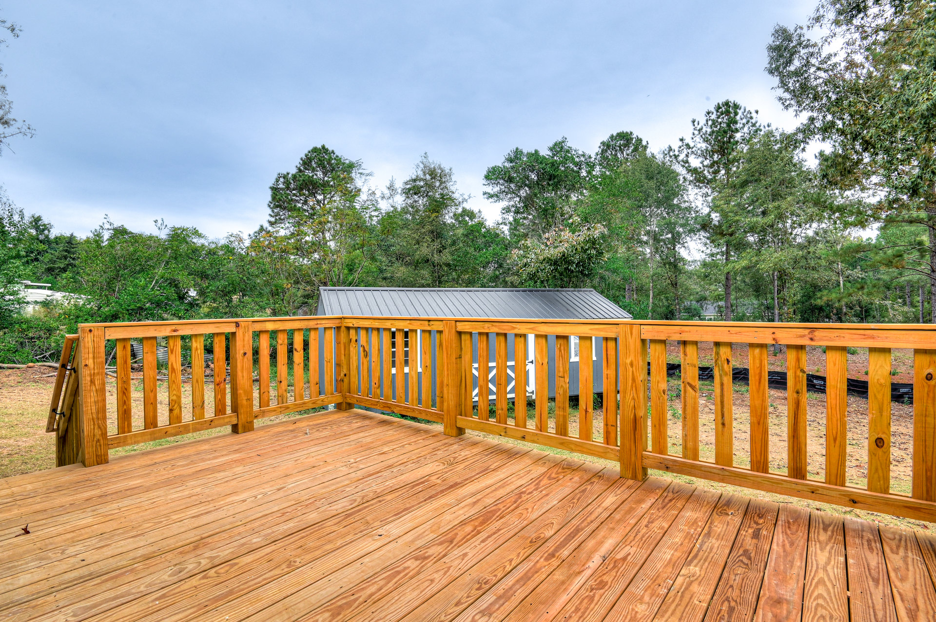 Wooden deck with horizontal plank flooring, sturdy wooden railing, and leafy trees beyond the fence under a partly cloudy sky
