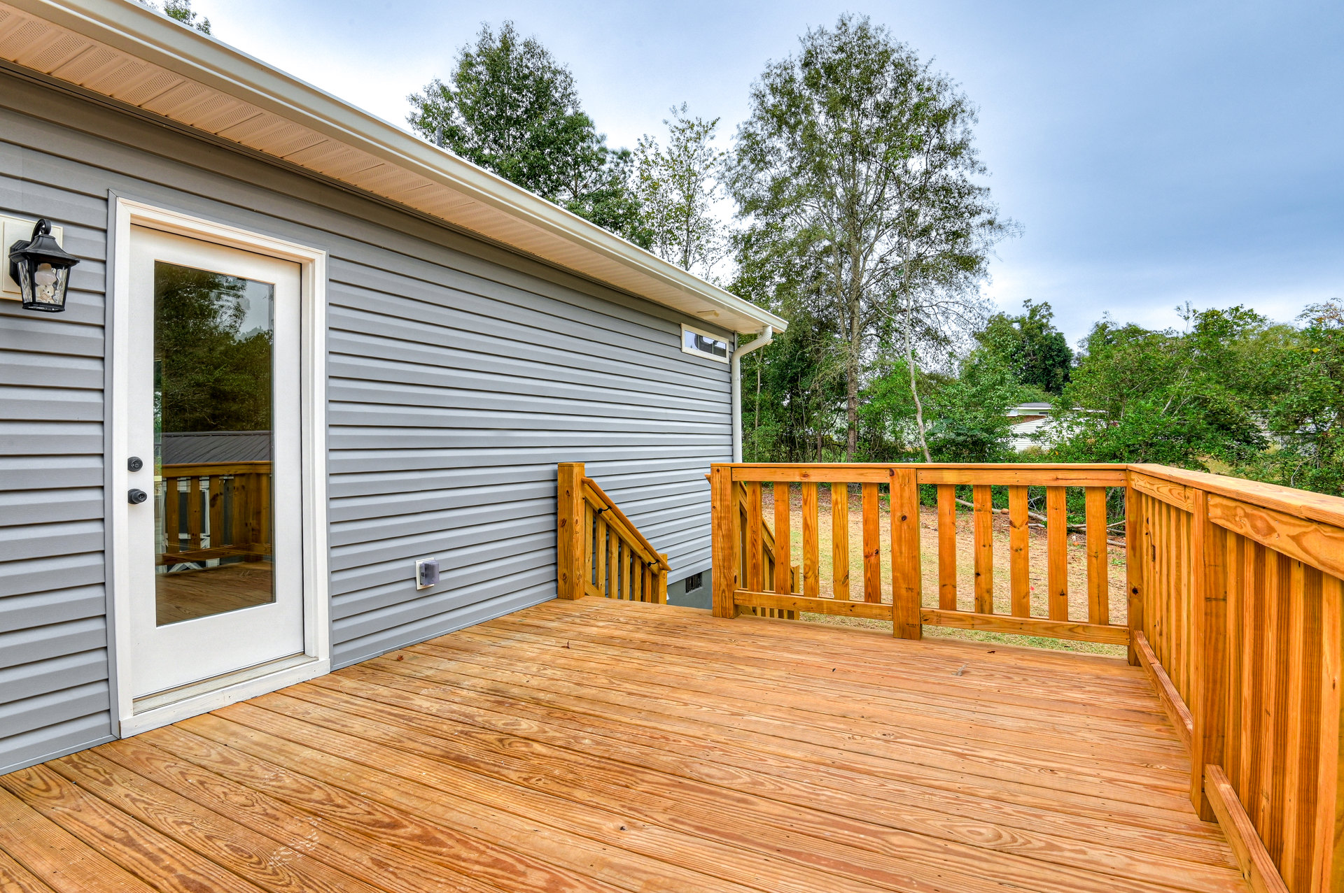 Wooden deck with plank flooring, wood railing, glass door, and leafy tree beside house exterior with light-colored siding