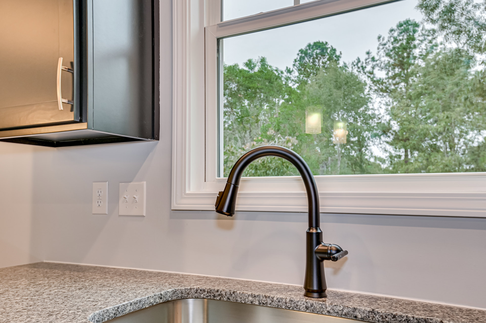 White kitchen sink with black faucet set beneath a window showing green trees, light switch with two toggles on wall, blurred pendant light, and door handle visible.