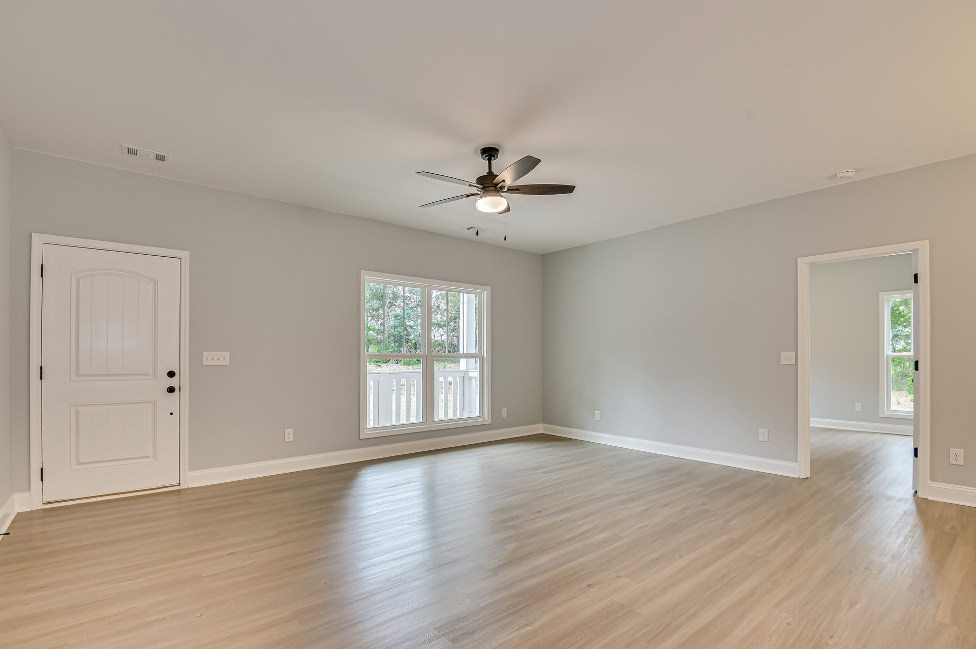 Ceiling fan with light fixture above wood laminate flooring, white walls, black-handled white door, large window overlooking trees