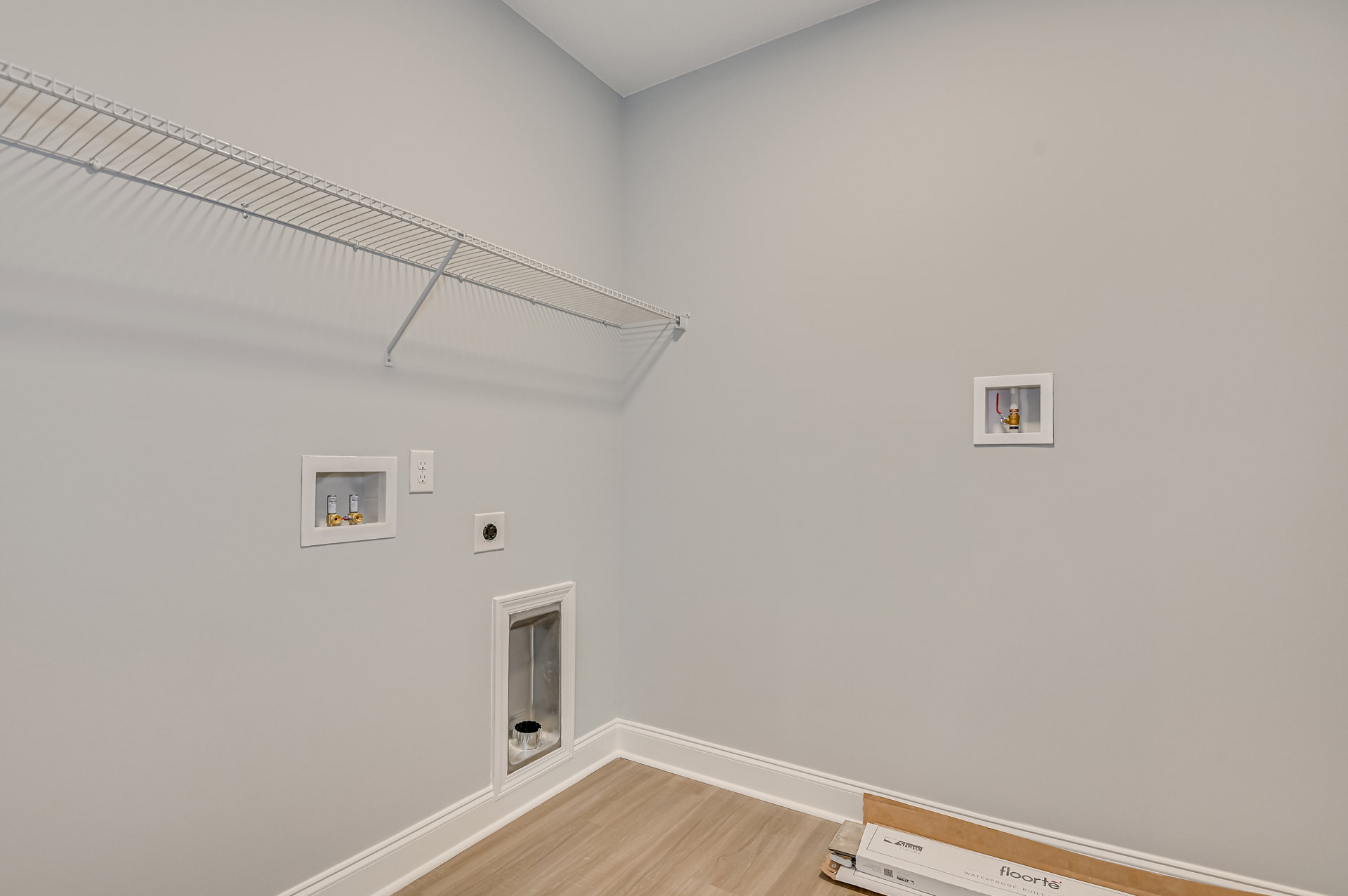 White plaster wall with built-in white shelves, white metal shelving unit, cardboard boxes on light floor, exposed pipes with red handle, and molding along ceiling.