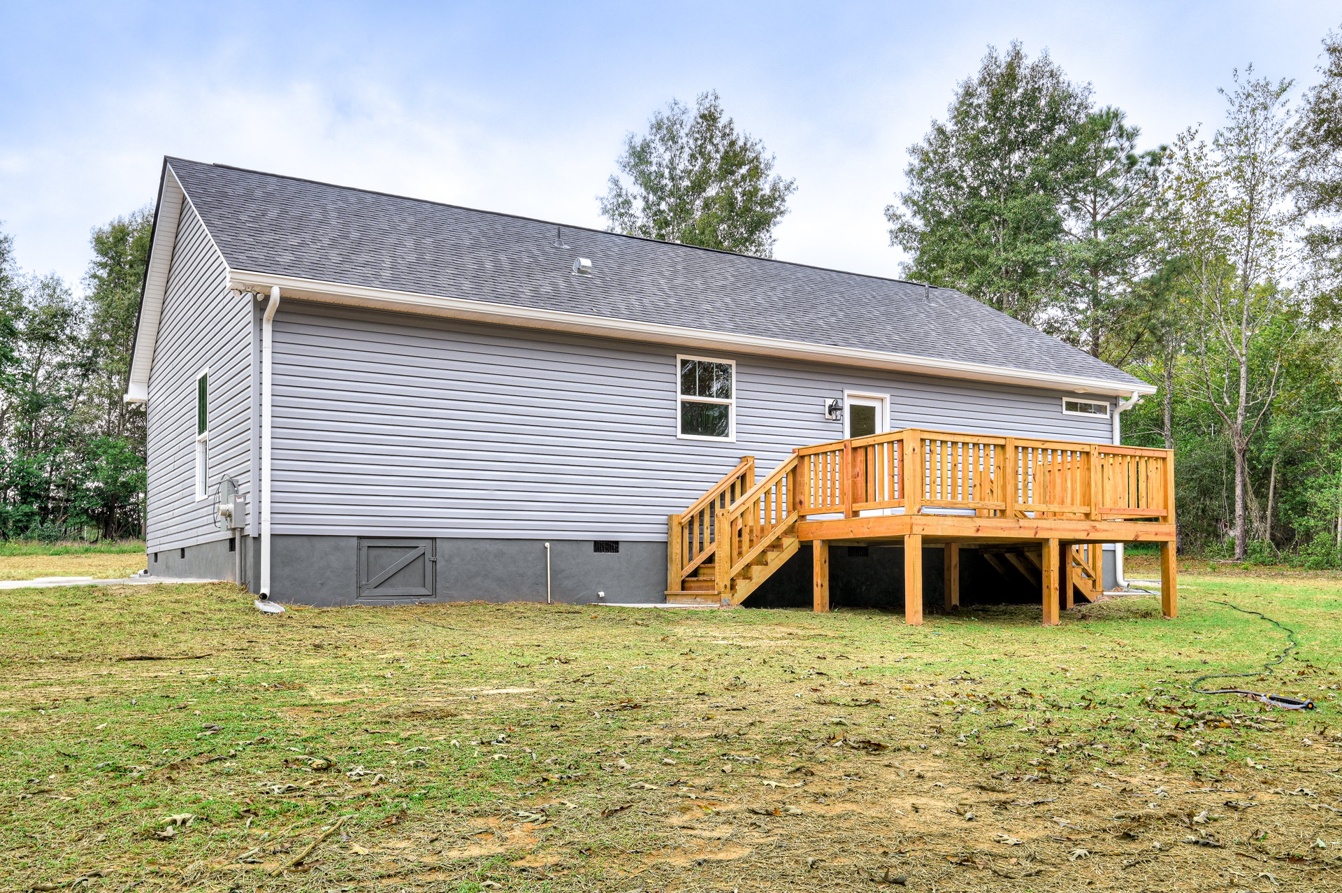 Two-story cottage with grey siding, white-framed windows, wooden deck and staircase, grassy lawn, and mature trees under a partly cloudy sky