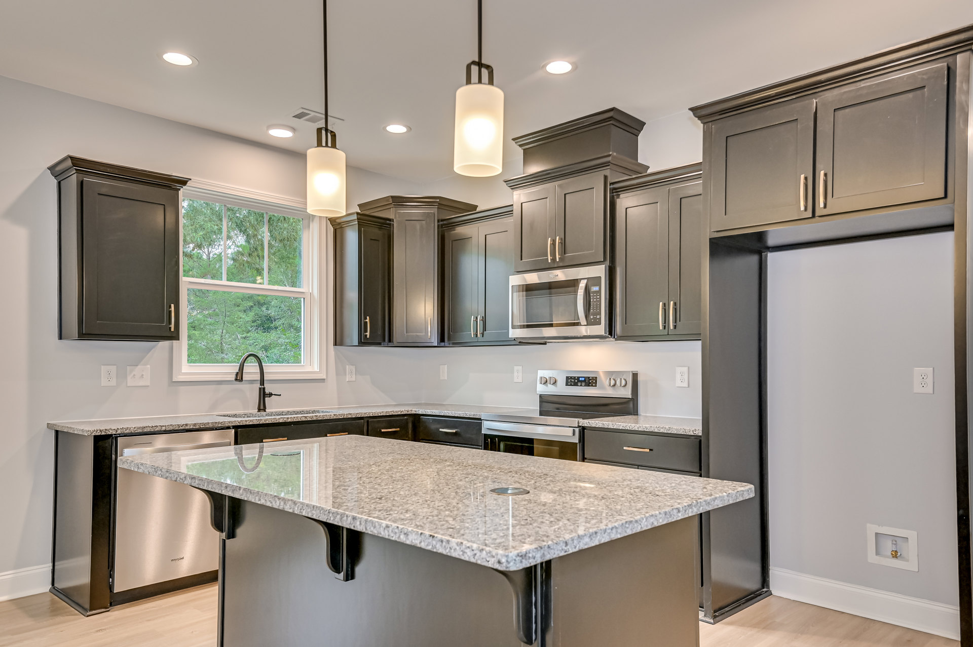 Marble kitchen island with built-in sink, silver and black countertops, glass door microwave, black door with silver handle, wall-mounted light fixture, white cabinetry, and