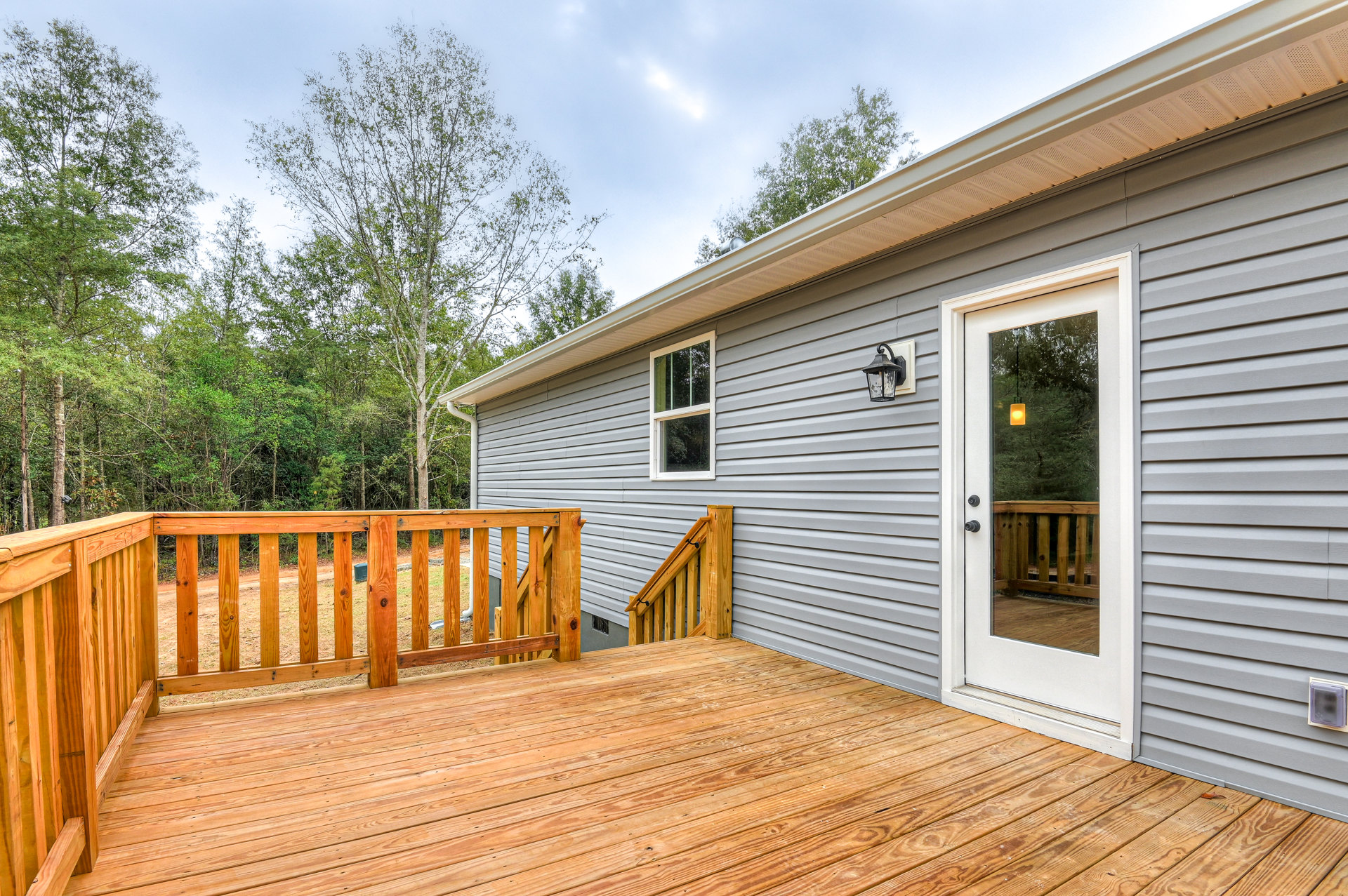 Wooden deck with horizontal wood railing, exterior door with light shining through, window with green curtain, light-colored siding, trees in background