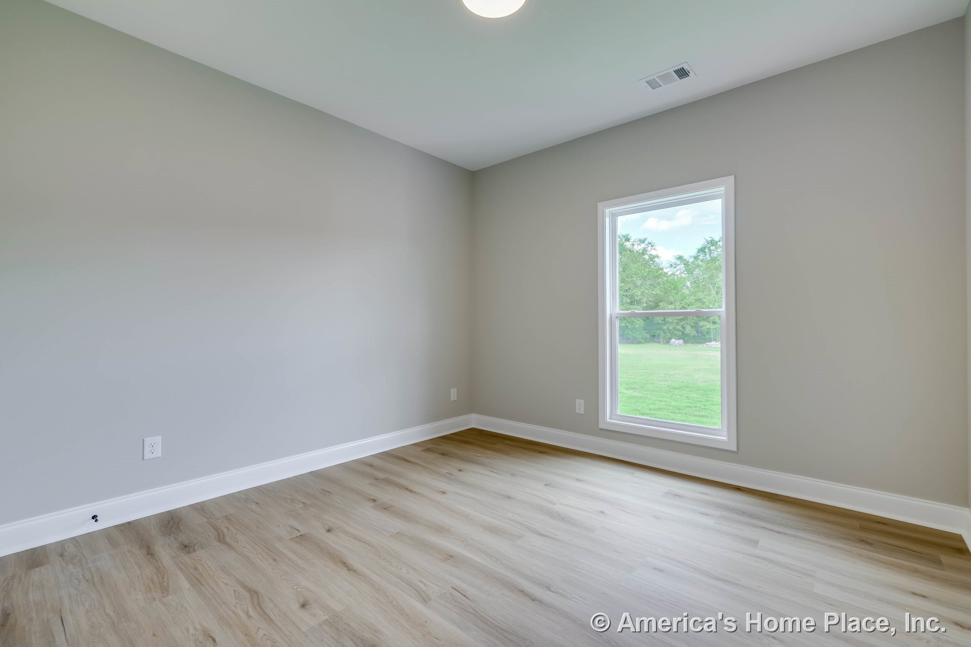 Rectangular bedroom with light gray walls, white baseboards, wood-look vinyl plank flooring, large single window with white trim, flush ceiling light fixture, and abundant natural