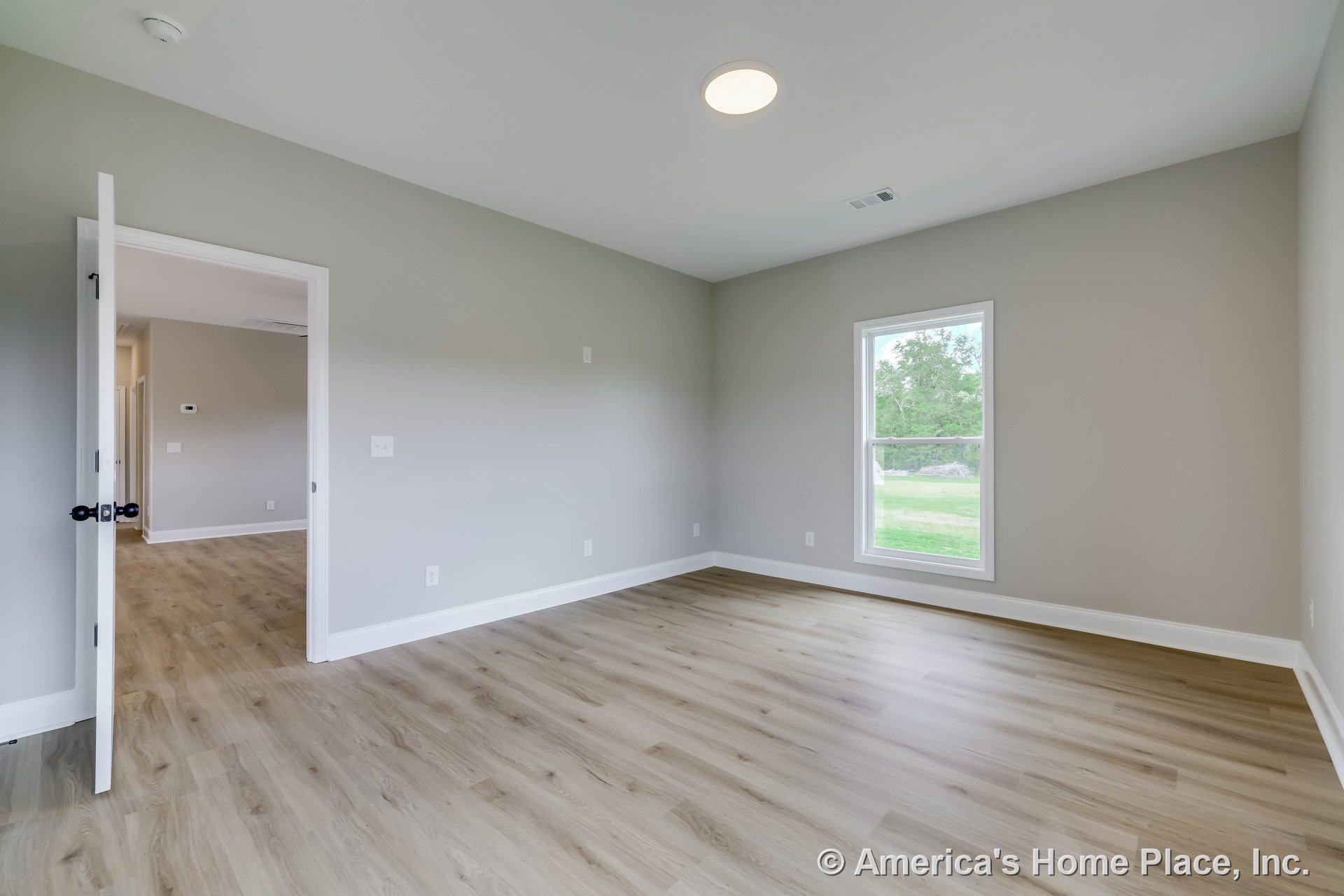 Bedroom with light wood-look flooring, neutral gray walls, single window framed in white trim, white baseboards, flush ceiling light fixture, and open white door.