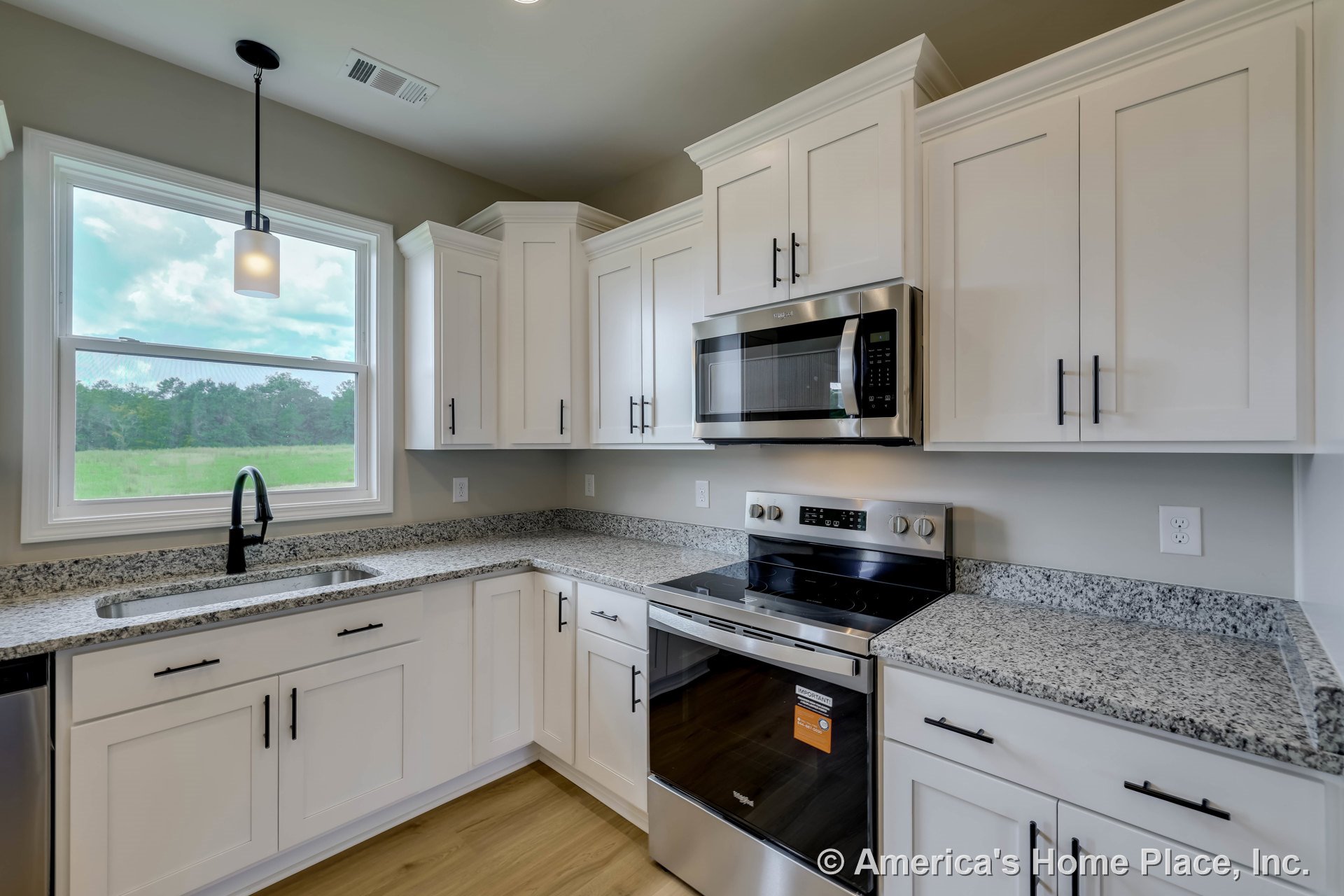 White shaker cabinets and granite countertops with stainless steel oven and microwave, large window above sink, modern pendant light fixture, and wood-look flooring.