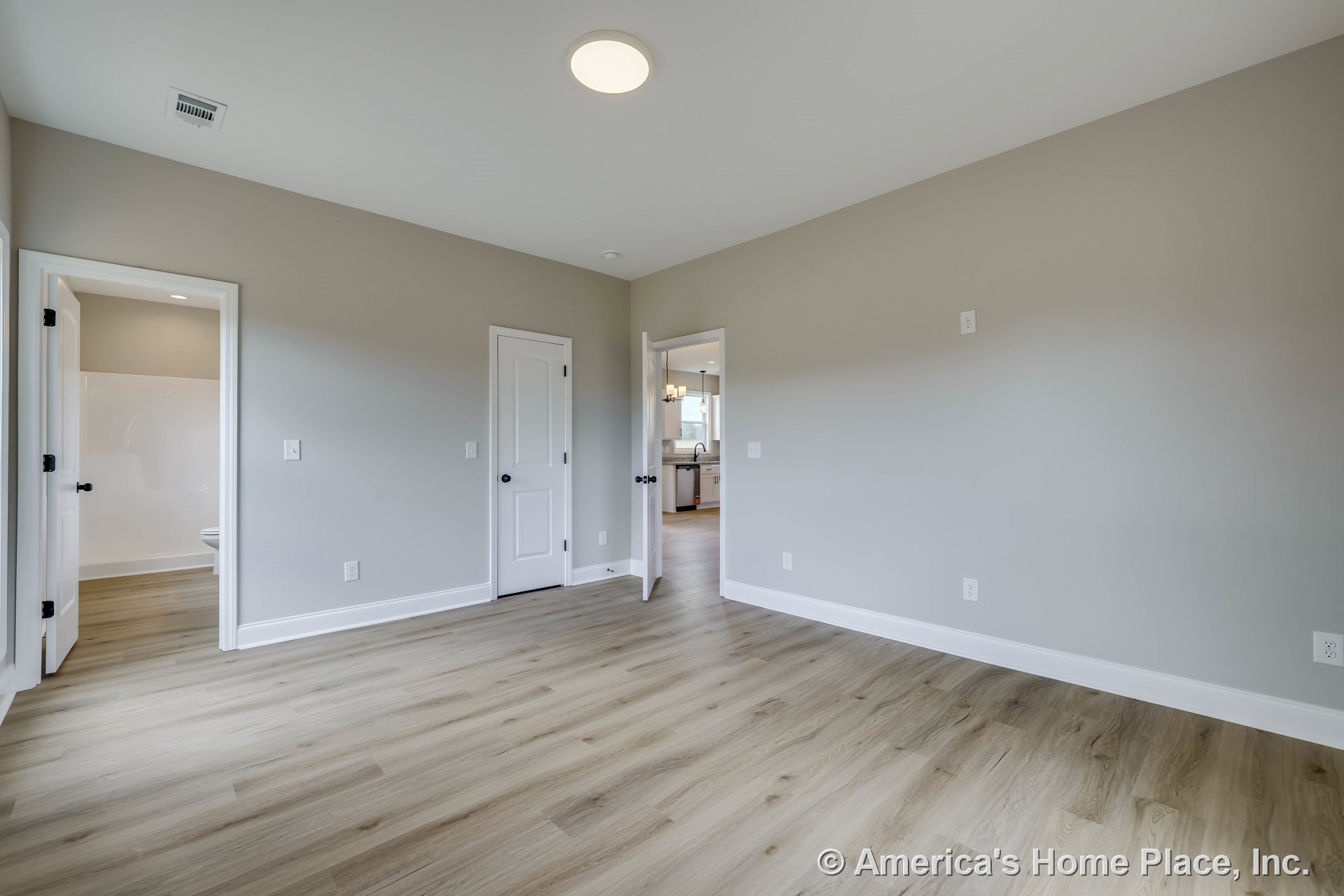 Bedroom with light wood plank flooring, neutral painted walls, white baseboard trim, flush mount ceiling light, two-panel interior doors, open doorway leading to bathroom with