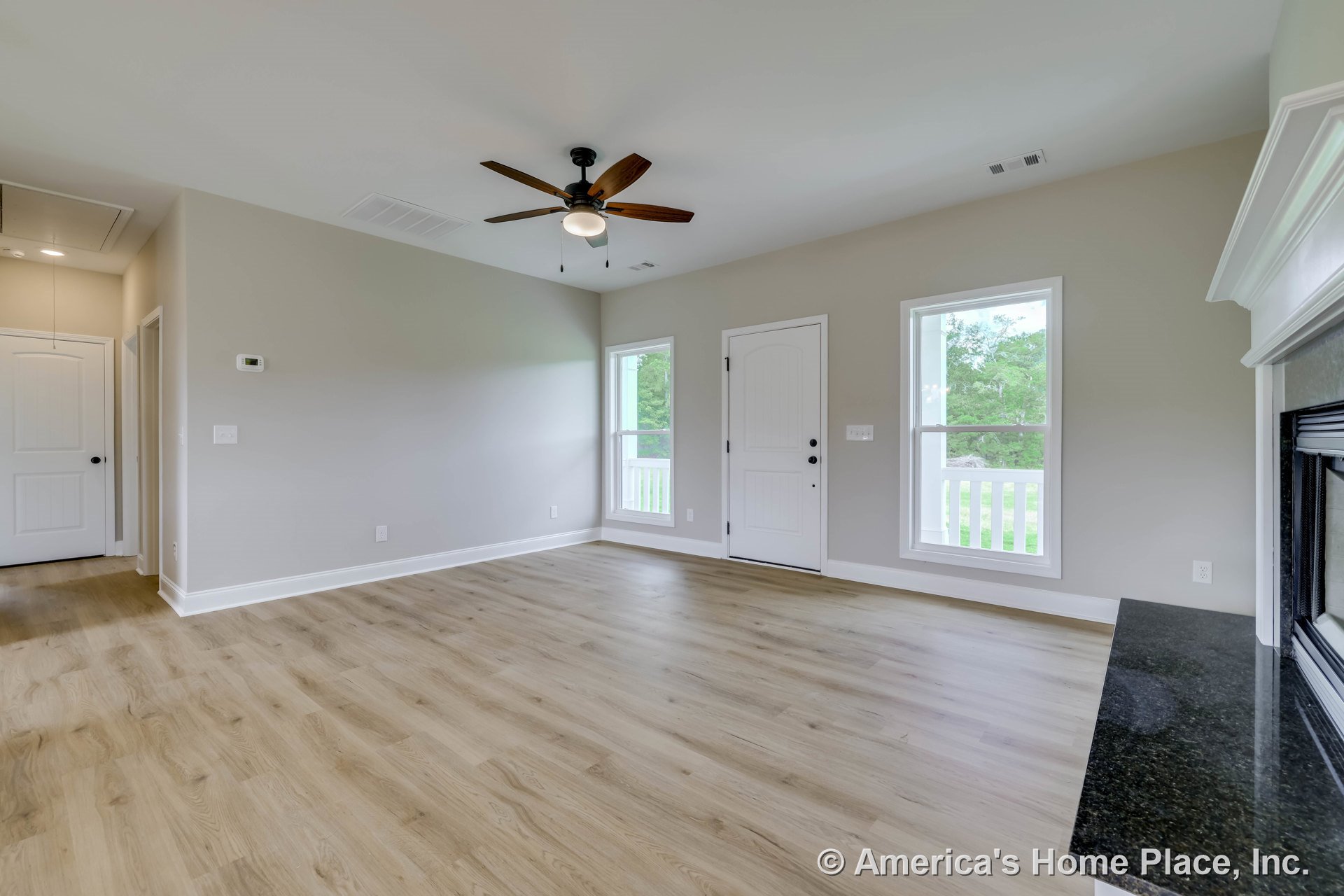 Living room with light wood plank flooring, white trim and baseboards, ceiling fan with integrated light, fireplace featuring a black stone mantel, two large windows, and a white