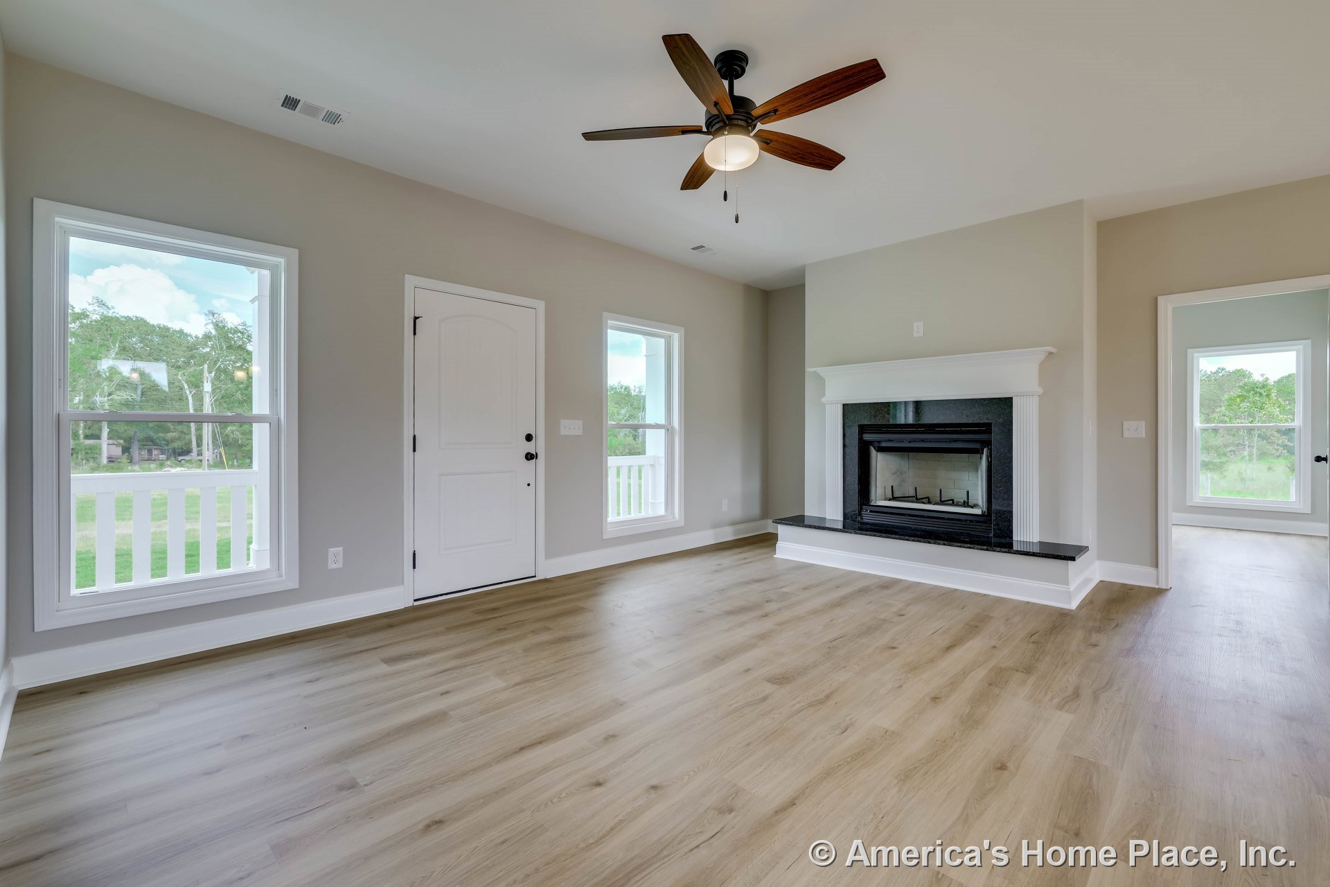 Central living room with black-surround fireplace and white trim, wood-look plank flooring, large white-framed windows, ceiling fan with light, white entry door, neutral wall