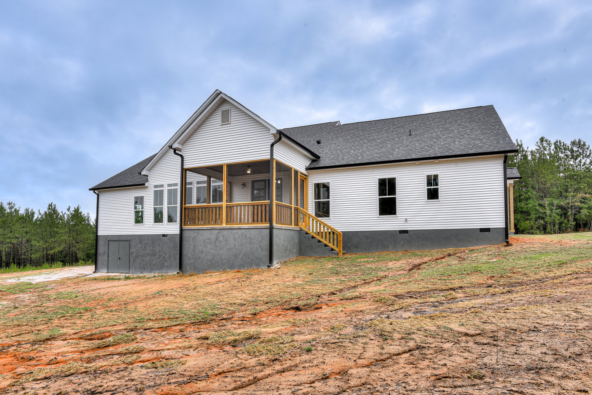 Two-story house with white-framed windows, covered front porch, concrete retaining wall and staircase leading up a grassy hill, surrounded by mature trees under a cloudy sky