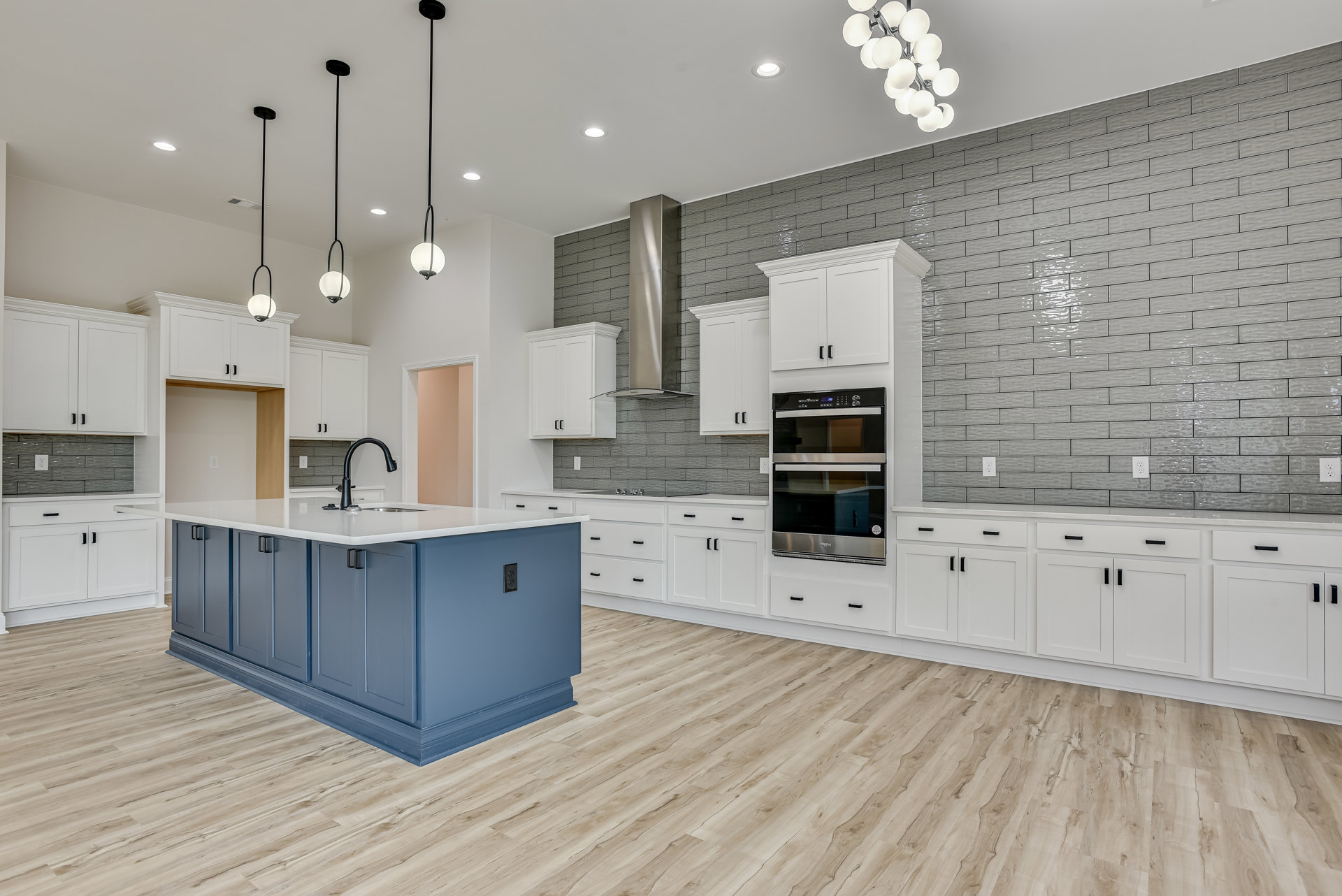 Blue kitchen island with built-in sink, white cabinets, black and silver oven, wood flooring, modern chandelier with white globes, blue baseboards, black support pole against white