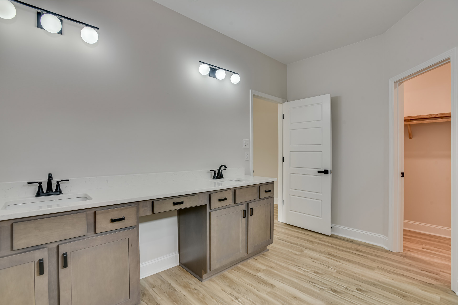 Bathroom with white double sink vanity, white countertop, wood flooring, white door with black handle, and modern light fixture featuring four round bulbs on a black metal bar.