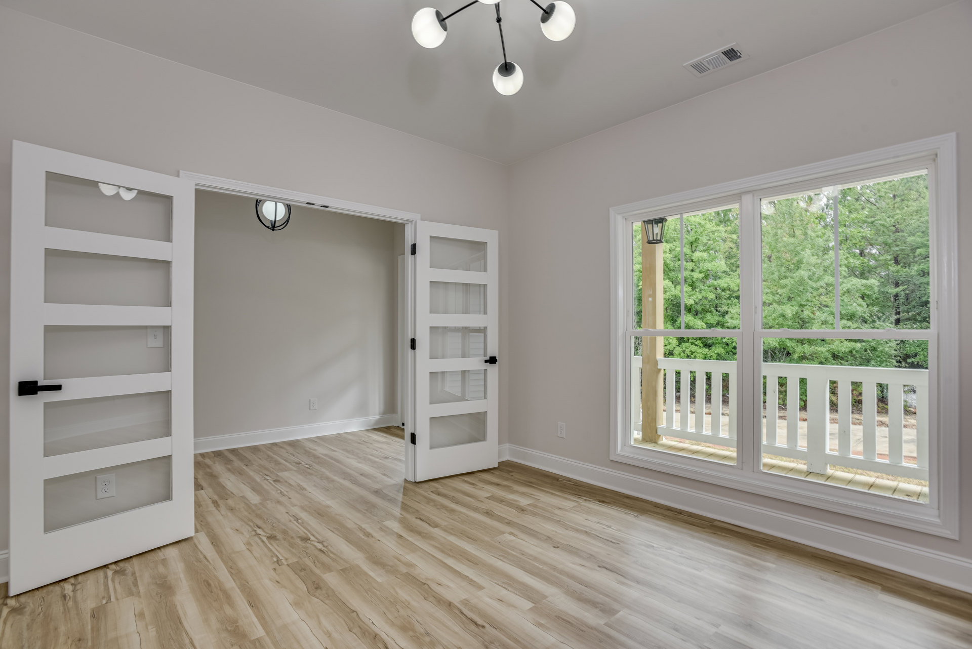 Wood floor room with two white doors featuring glass panels, large window showing trees outside, and a modern chandelier suspended from the ceiling.