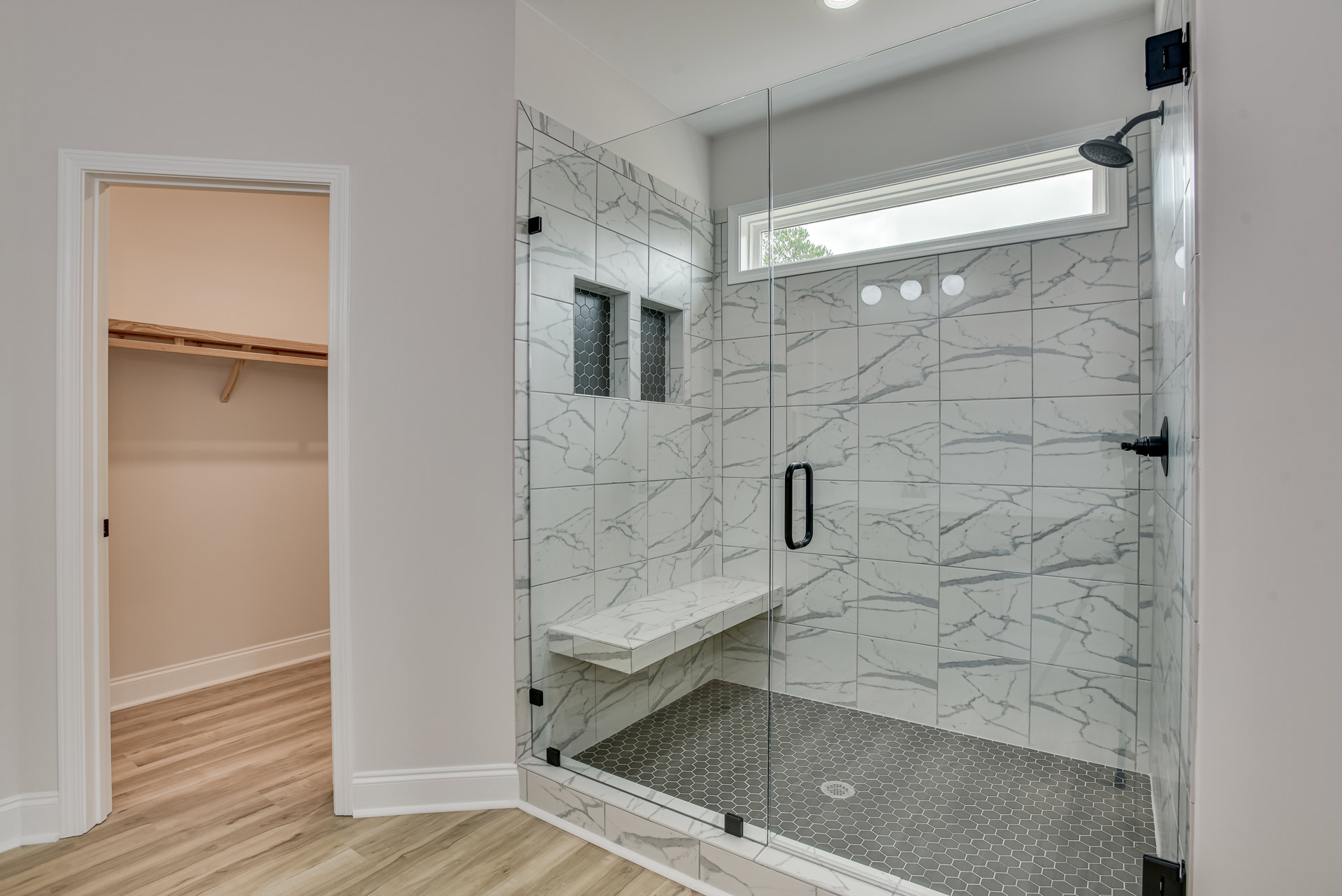 Glass shower enclosure with a white marble bench, black and white marble tile walls, and a window providing natural light