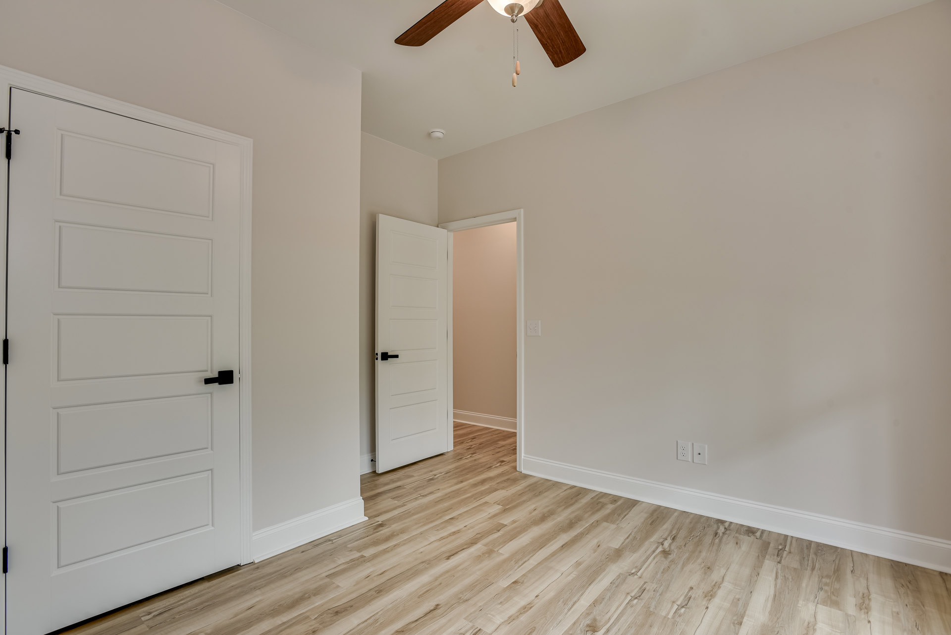 Wood floor and white walls, ceiling fan with light fixture, white door with black handle, simple modern interior
