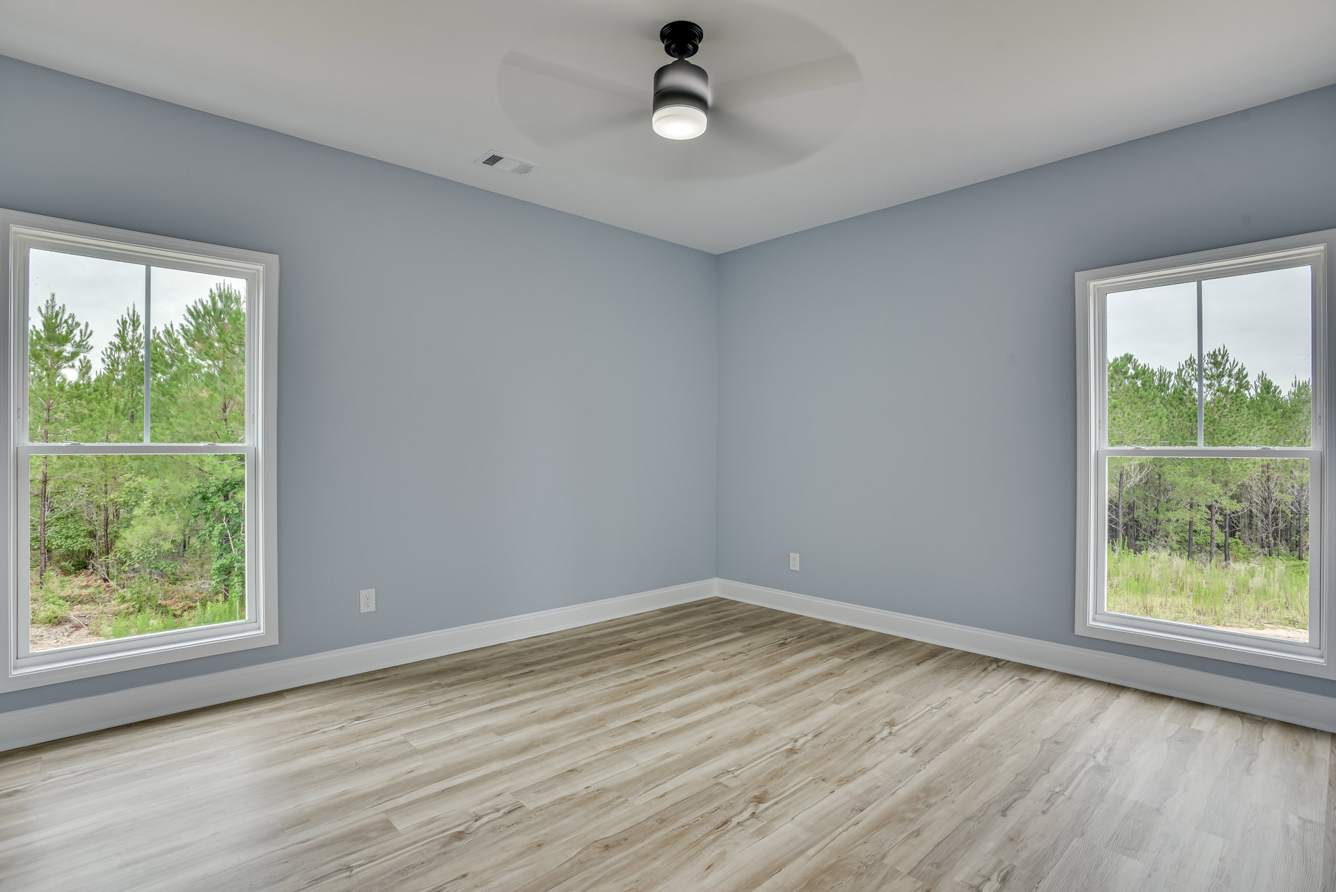 Wood flooring in a bright room with large windows, ceiling fan with light fixture, and views of trees outside