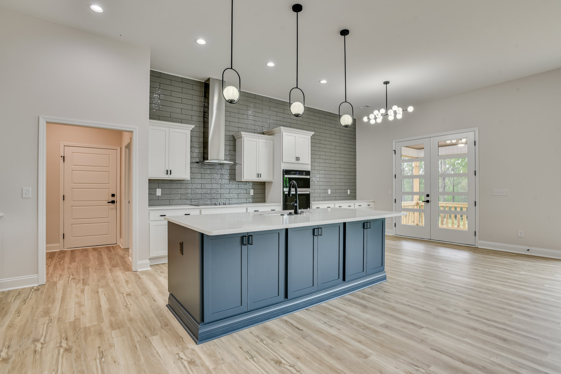 Spacious kitchen featuring a large island with white countertop, tile flooring, white cabinetry, stainless steel sink, glass-panel double doors, and black-handled white door.
