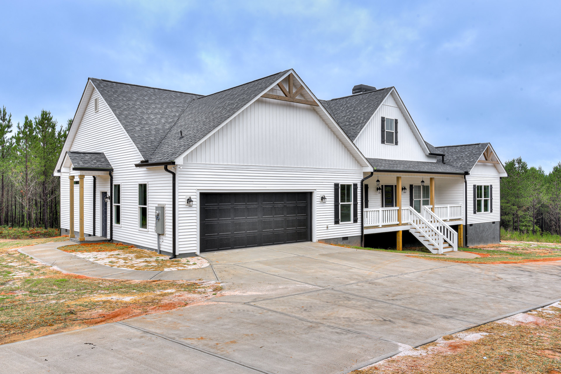 Black garage door with white trim, white staircase leading to entry, white-framed windows, driveway, surrounded by trees and cloudy sky