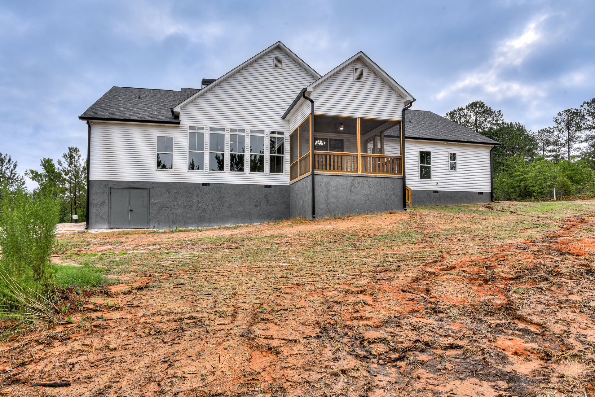 Two-story house with covered front porch, grey door, white-framed windows, concrete retaining wall, expansive grassy yard, mature trees, and cloudy sky