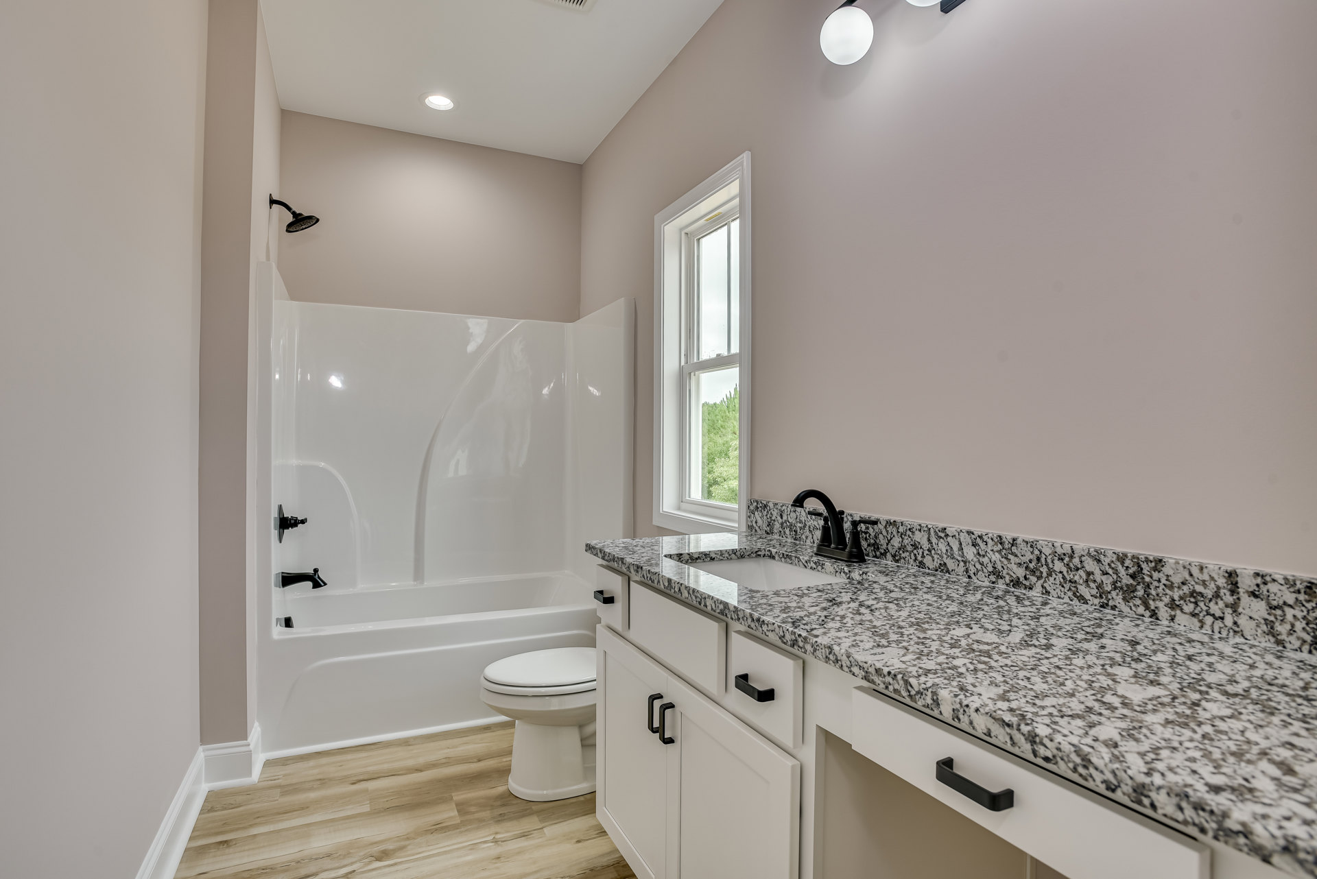 Modern bathroom with white porcelain sink, glass-enclosed shower, white toilet, light gray tile flooring, wood vanity, and a window with a green plant on the sill