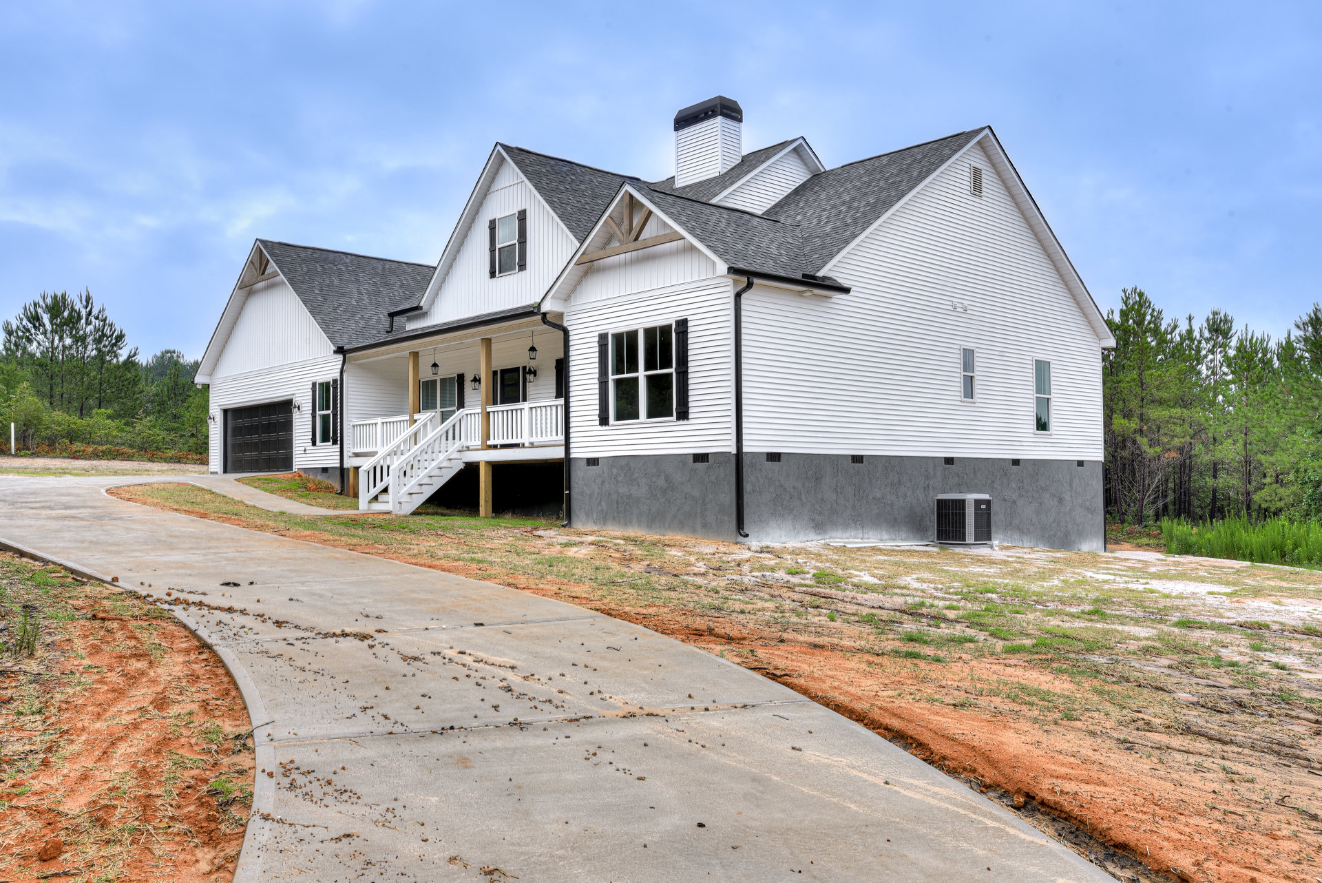 Two-story custom home with white siding, covered porch, brick chimney, concrete driveway, outdoor staircase, large windows, and landscaped front yard with trees and shrubs.