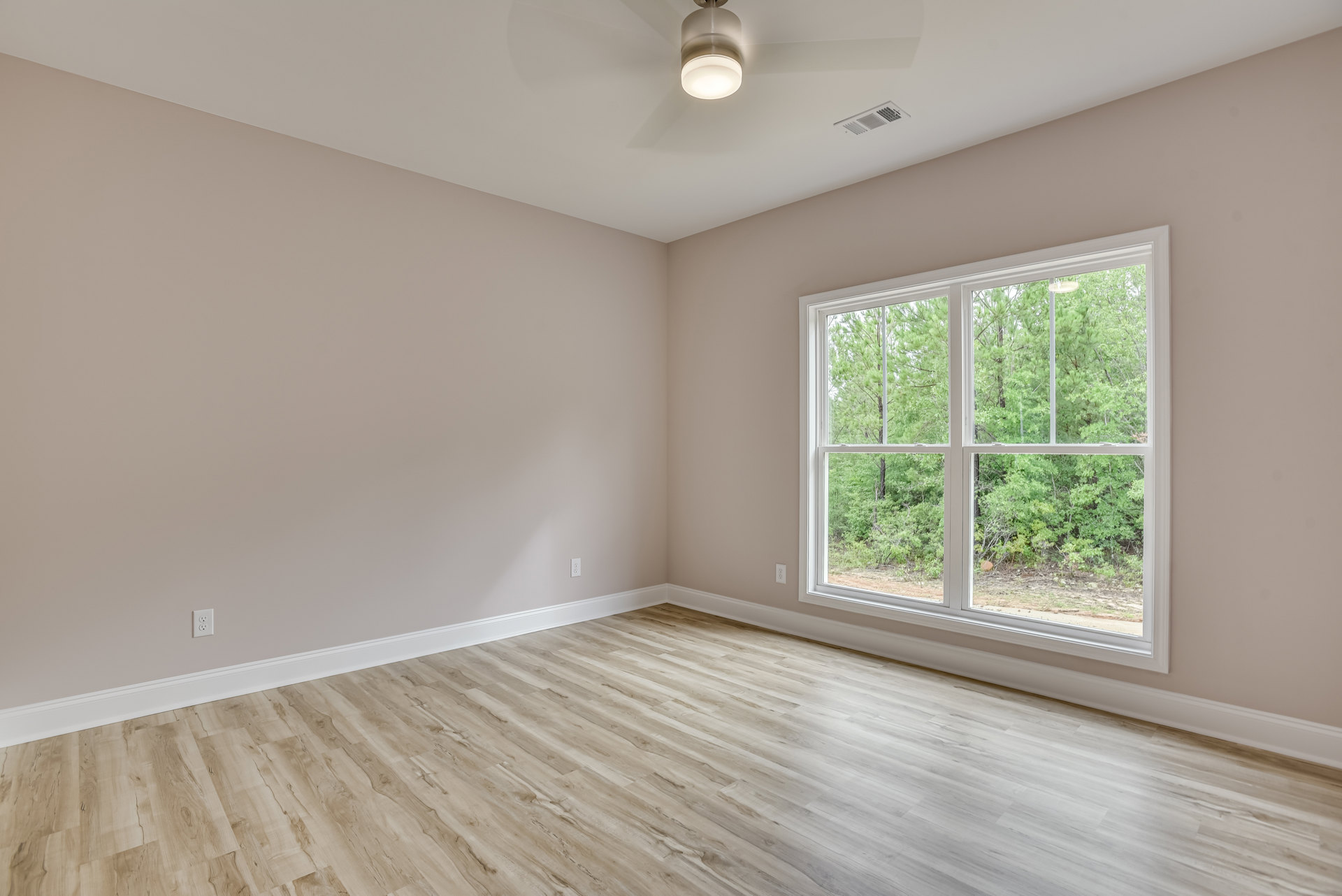 Sunlit room with large window overlooking trees, light wood flooring, white plaster walls, ceiling vent, and modern ceiling light fixture