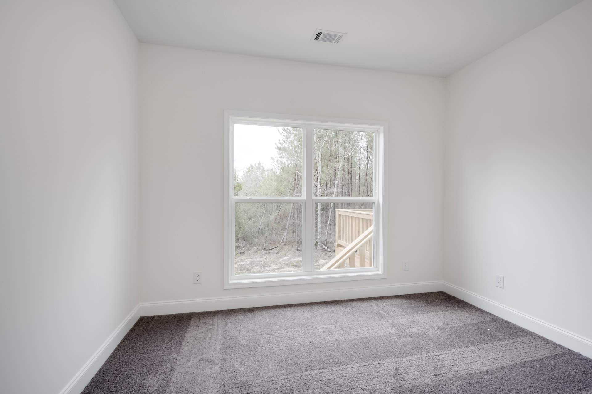 Carpeted bedroom with large window overlooking trees and wooden deck, white plaster walls, ceiling vent visible