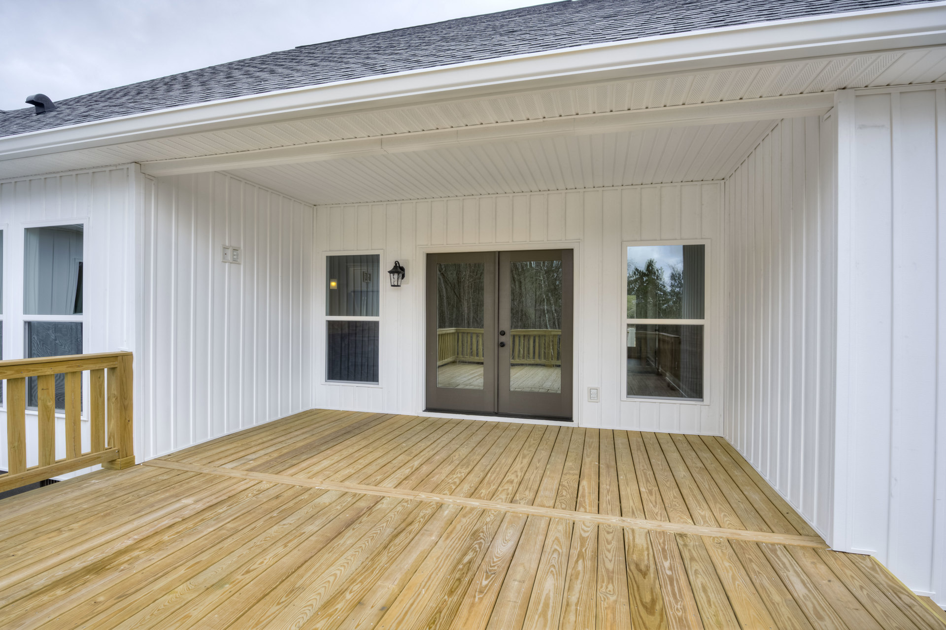 White siding house with glass double doors opening onto a wooden deck with railing, large window reflecting trees, and gabled roof.