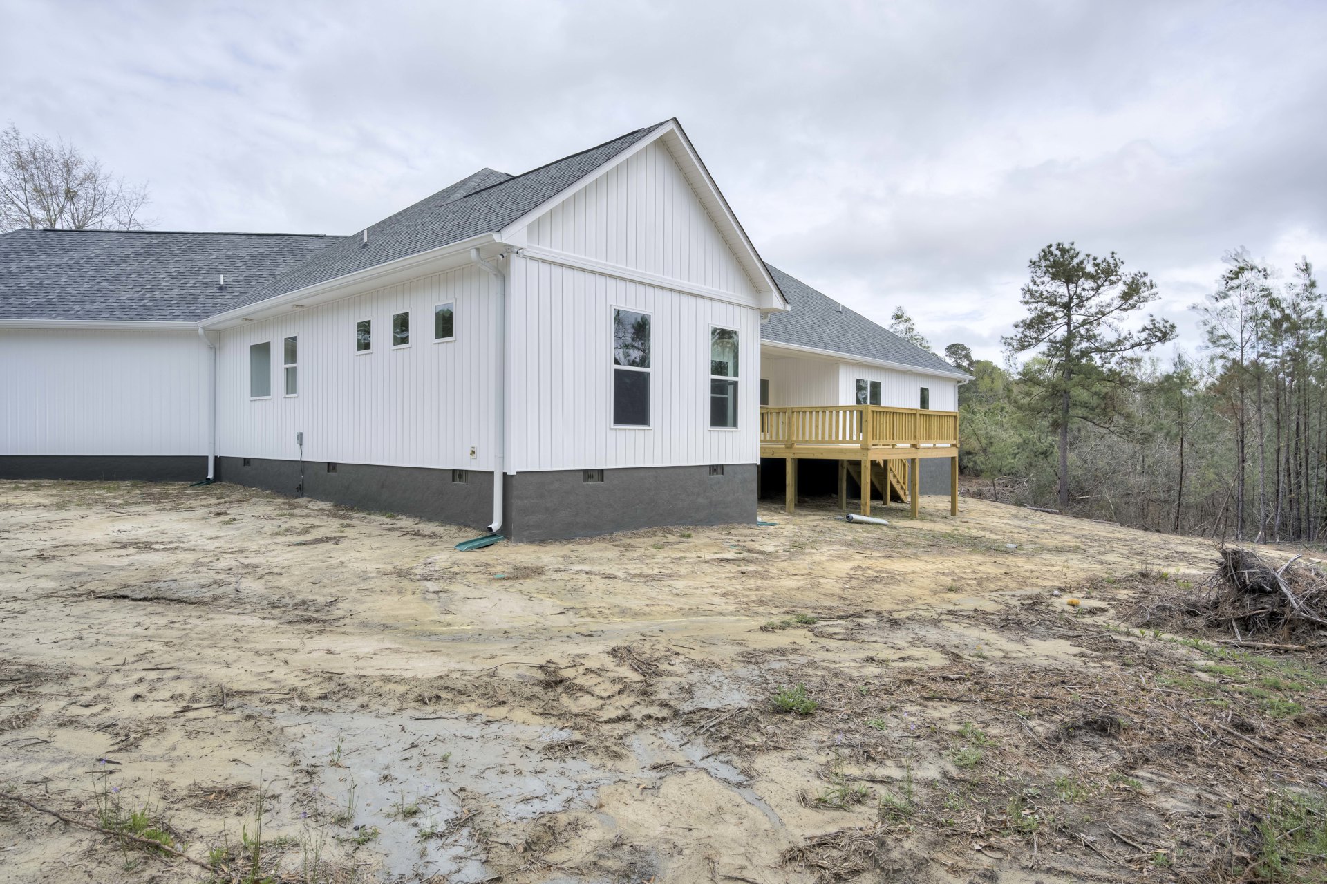 White house with black roof, wooden deck with stairs, large windows reflecting trees, dirt yard, detached garage, tall trees in background.